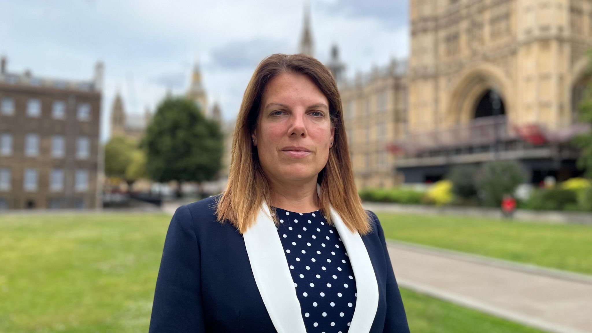 An image of Caroline Nokes MP, who is wearing a navy top with white polka dots and a navy jacket with a white lapel, at College Green