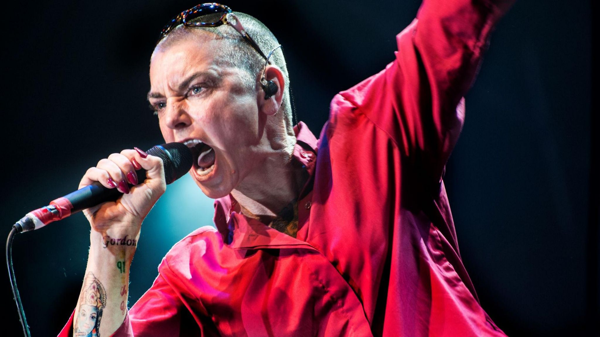 Sinéad O'Connor wears a pink button up shirt and sunglasses on top of her shaved head while singing into a wired microphone