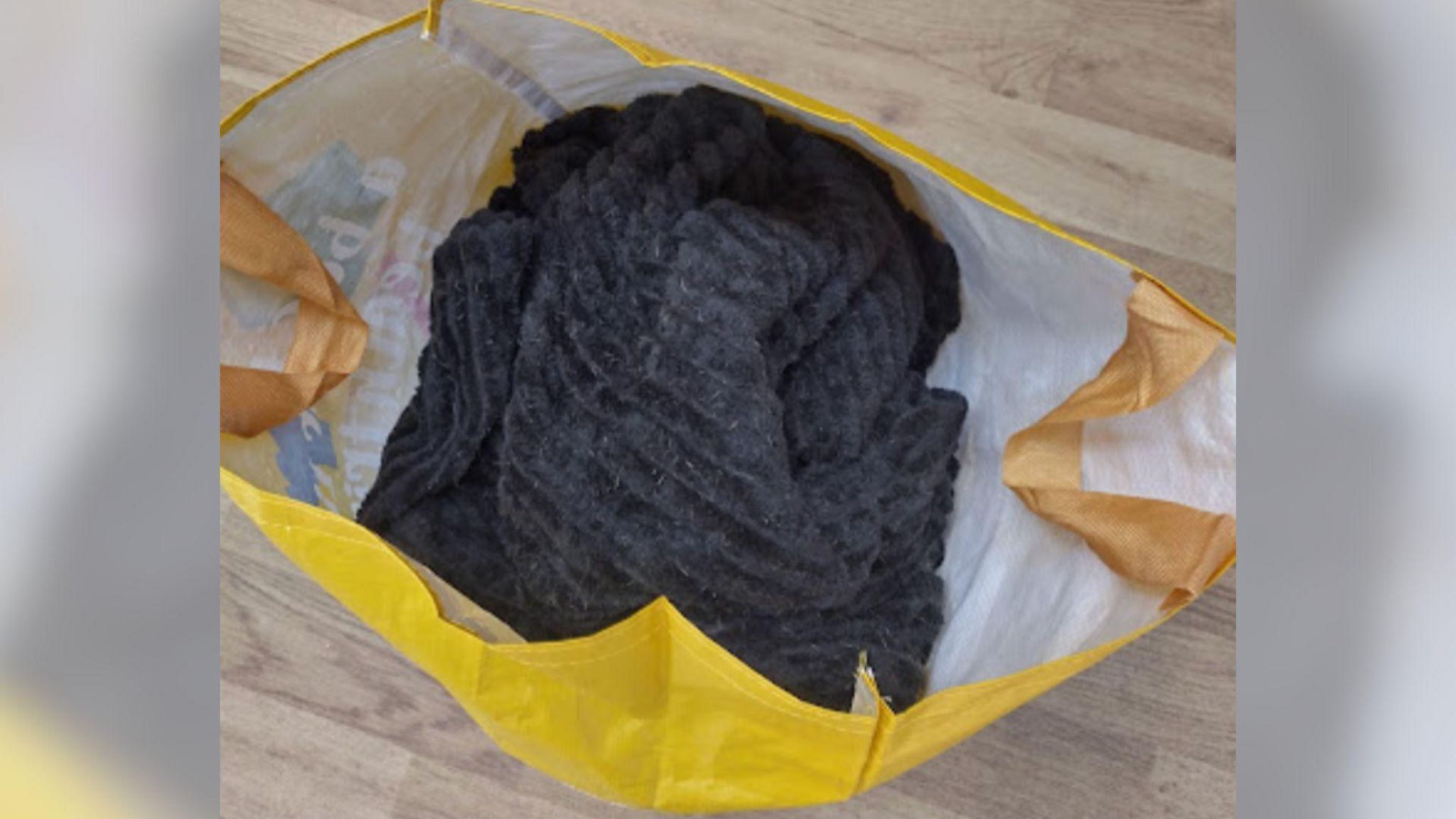 A view from above a yellow shopping bag. The inside of the bag is white..The bag has orange handles and is on light wood flooring. Inside the bag is a black blanket