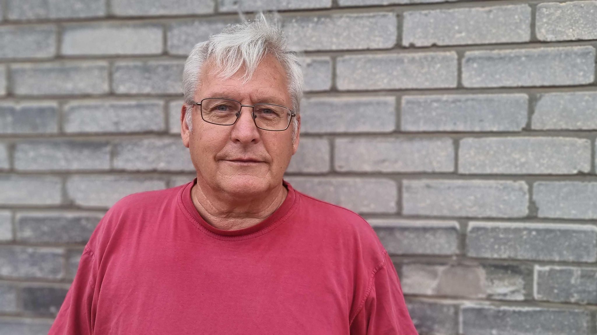 A head and shoulder shot of Prof David Martill, taken against a grey brick wall. He has short, white hair, and wears thin-rimmed rectangular glasses and a red T-shirt