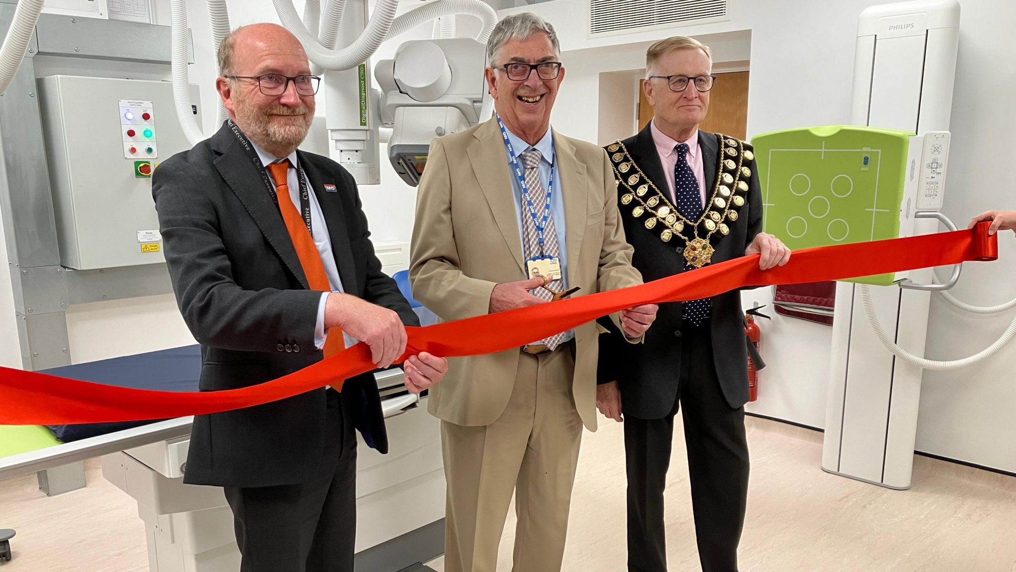 Three men in suits stand behind a red ribbon in front of the new X-ray machine