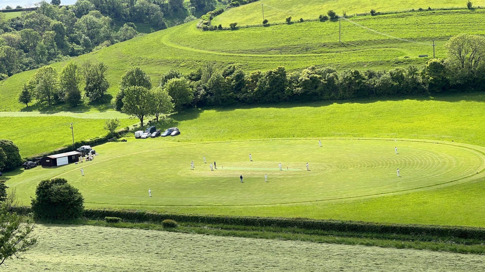An elevated view of a cricket field set in a rural landscape of trees and fields