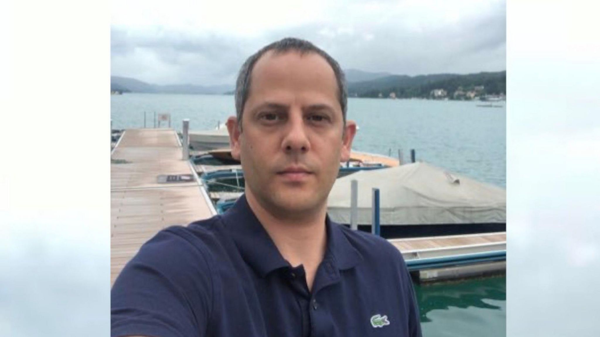 Eddy Eliaz is pictured looking straight to camera. He is wearing a dark blue T-shirt and is standing on a jetty, which appears to lead into a lake. There are speed boats moored next to the jetty and hills can be seen in the distance. Mr Eliaz has dark eyes and dark, short hair.