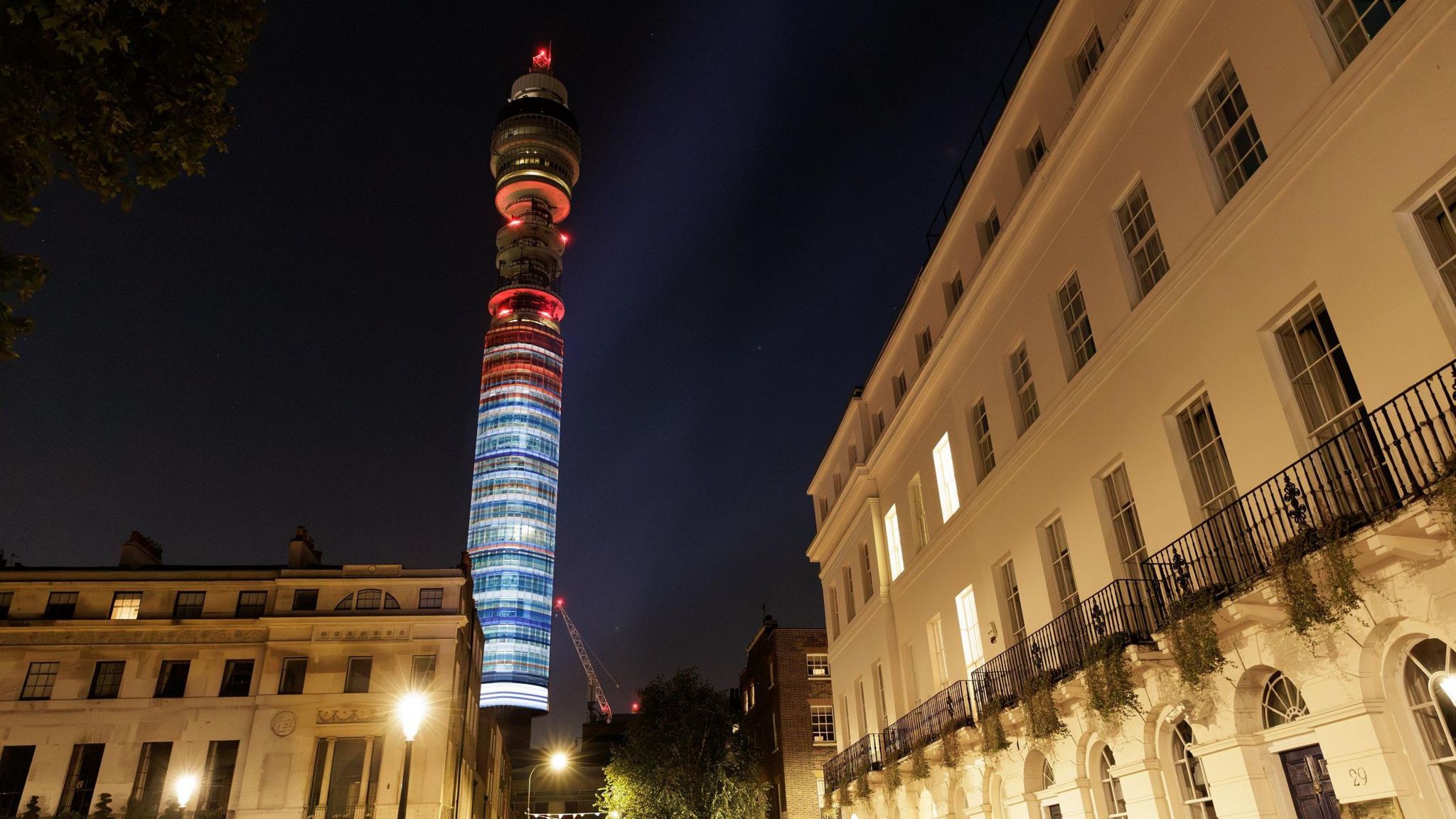 The blue-to-red gradient projected sideways onto a tall thin tower at night, with buildings lit by streetlamps in the foreground