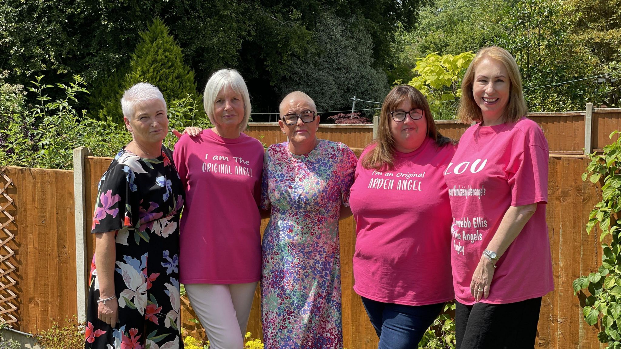 Nickie and Tracie with some of the Arden Angels. Three of the women wear pink T-shirts with white letters on them related to being Arden Angels while Nickie, on the left, wears a floral dress as does Ms Mills in the centre. They stand in a back garden in front of fence panels