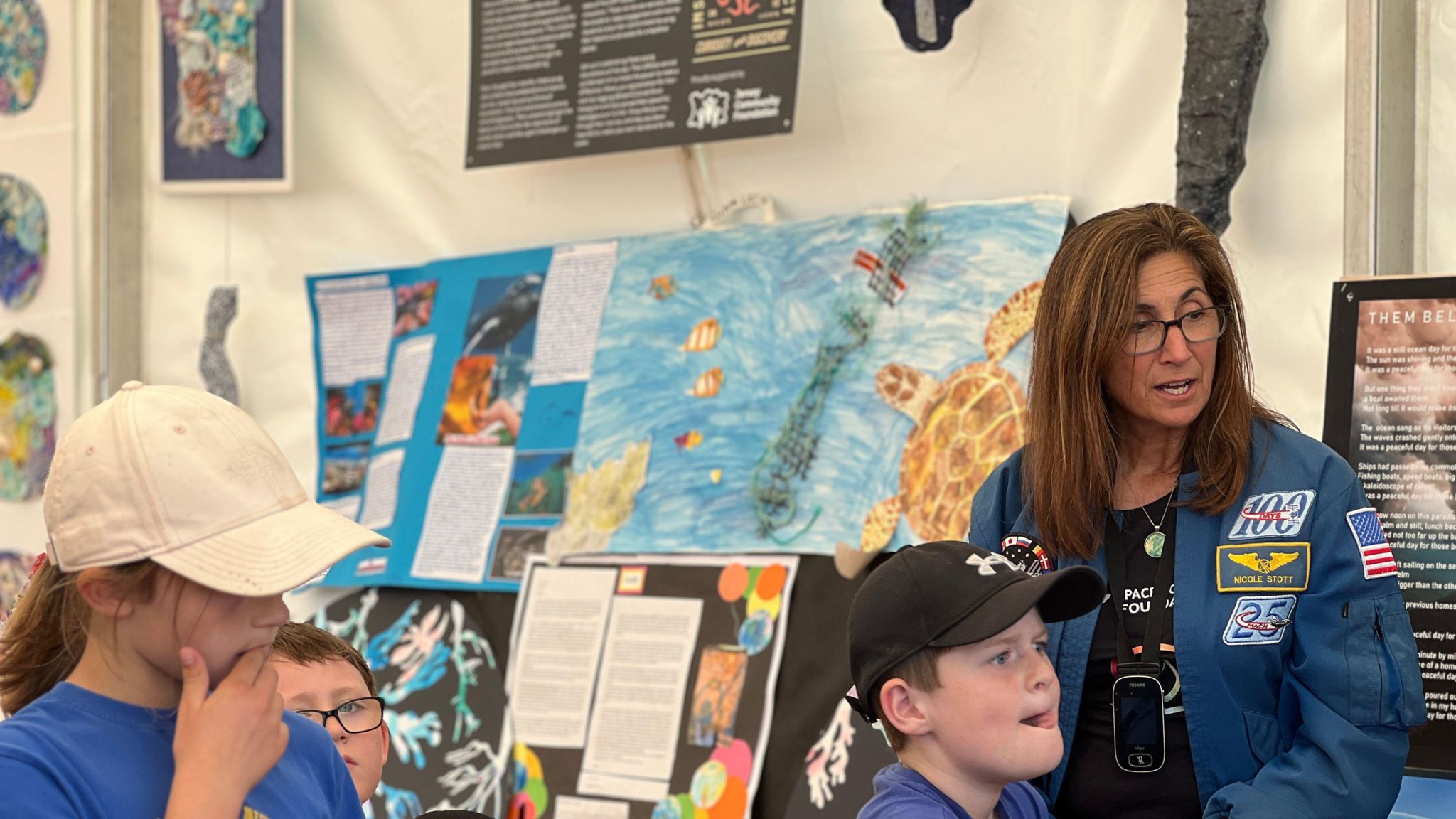 Nicole Stott is wearing a special astronaut jacket and sits next to some sea artwork and speaks to children sitting in front of her