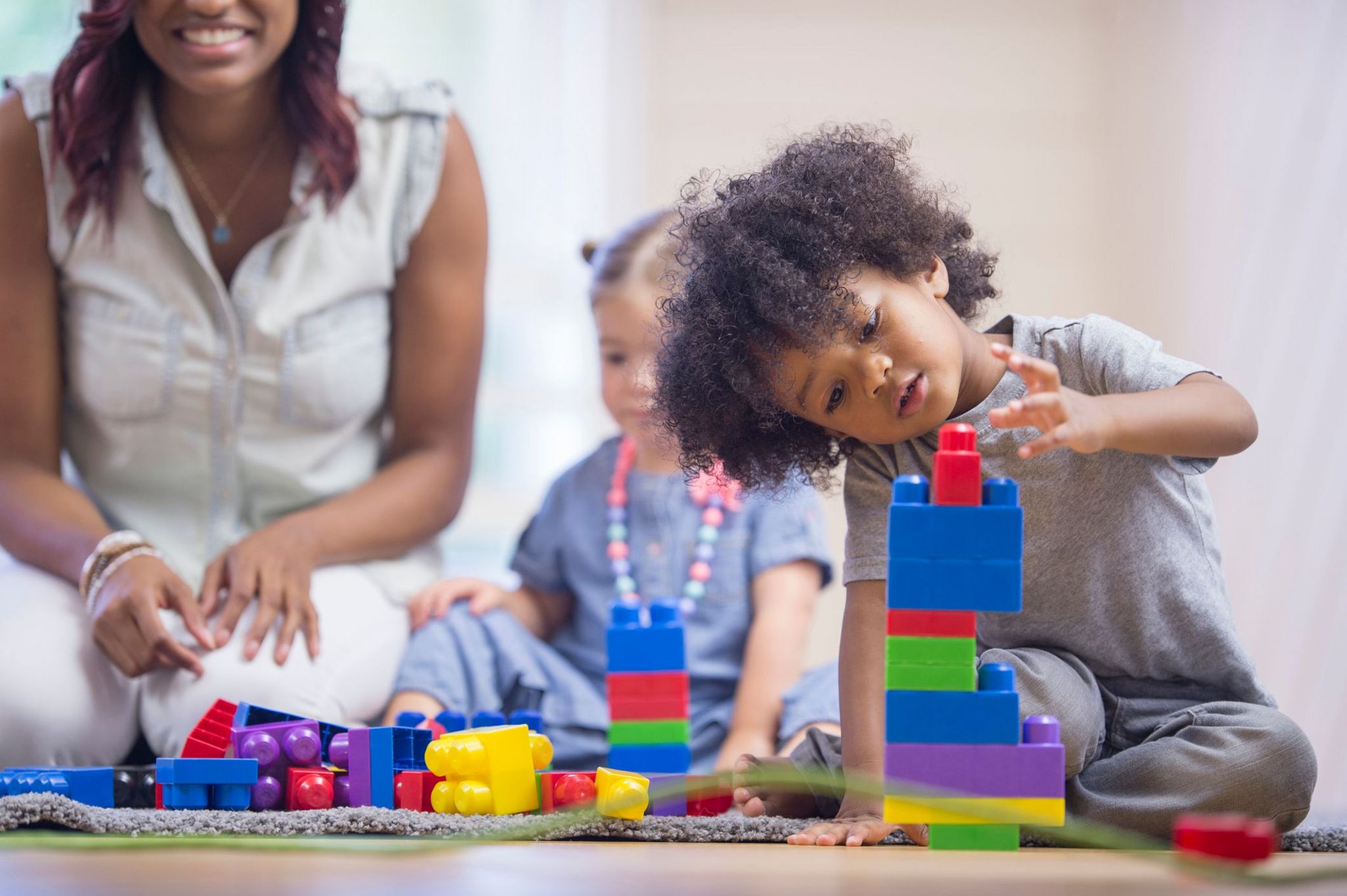 A stock image showing a young boy in the foreground playing with large stackable lego-type bricks. A young girl and a woman can be seen in the background also on the playmat.