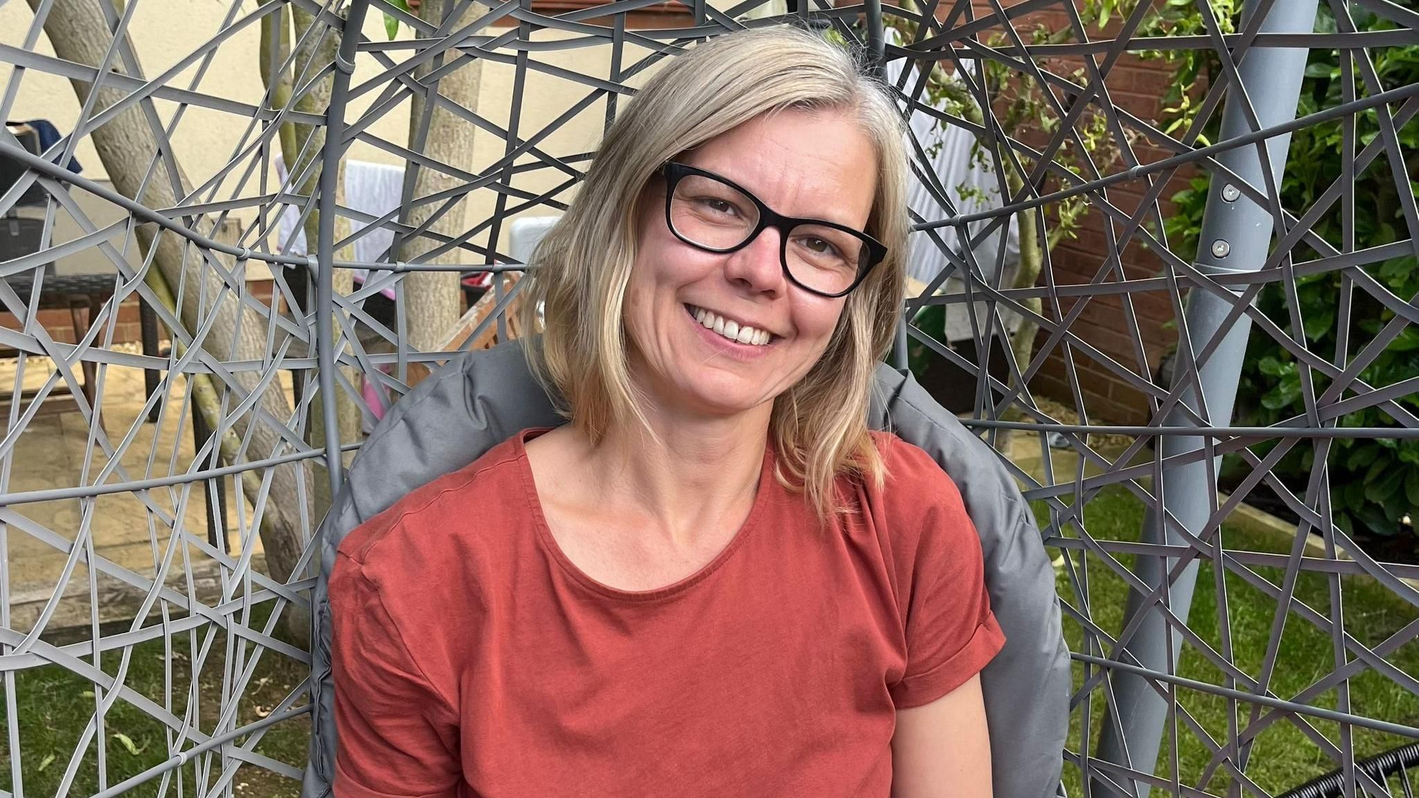 A lady in a red top with a blonde bob sitting outside looking at the camera 