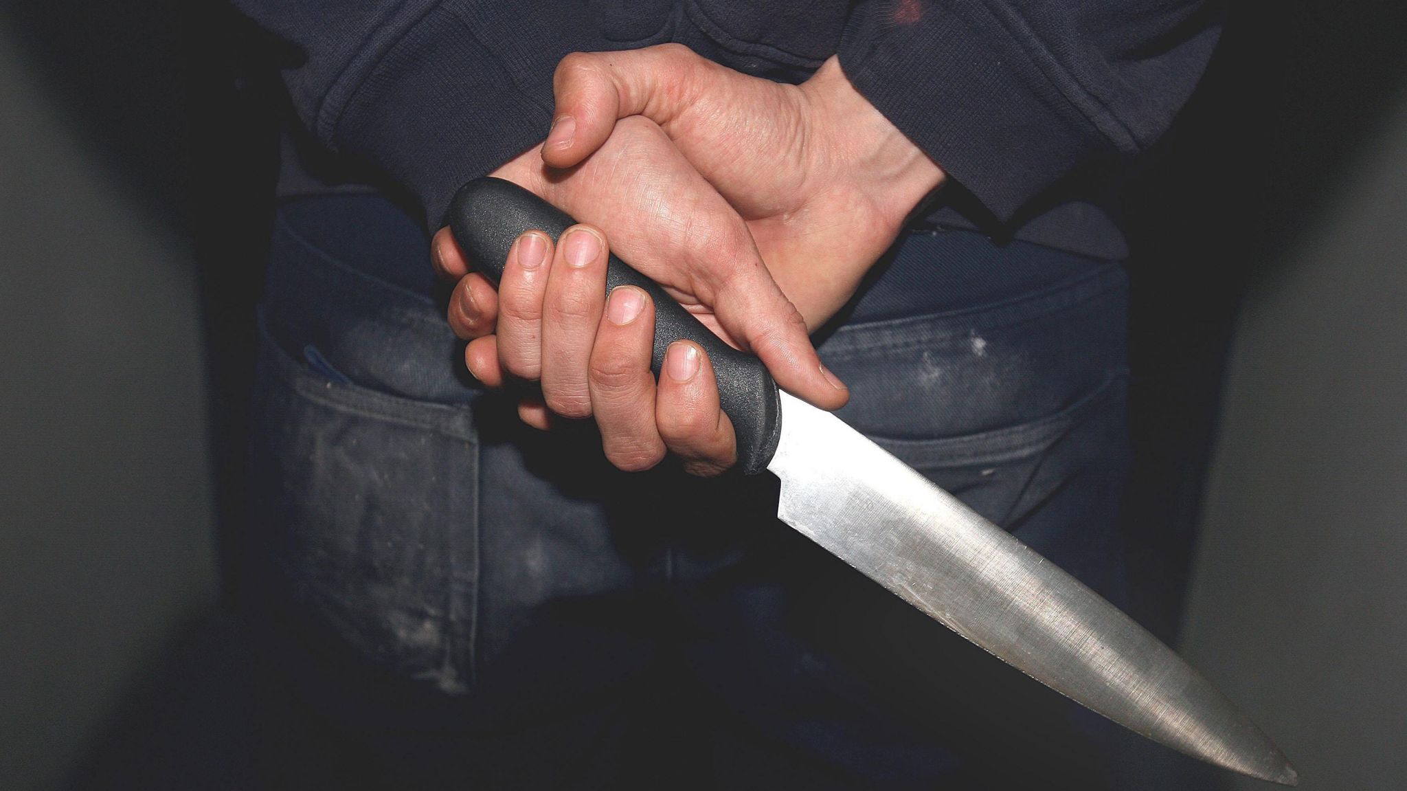 A young person stands with their hands behind their back holding a knife