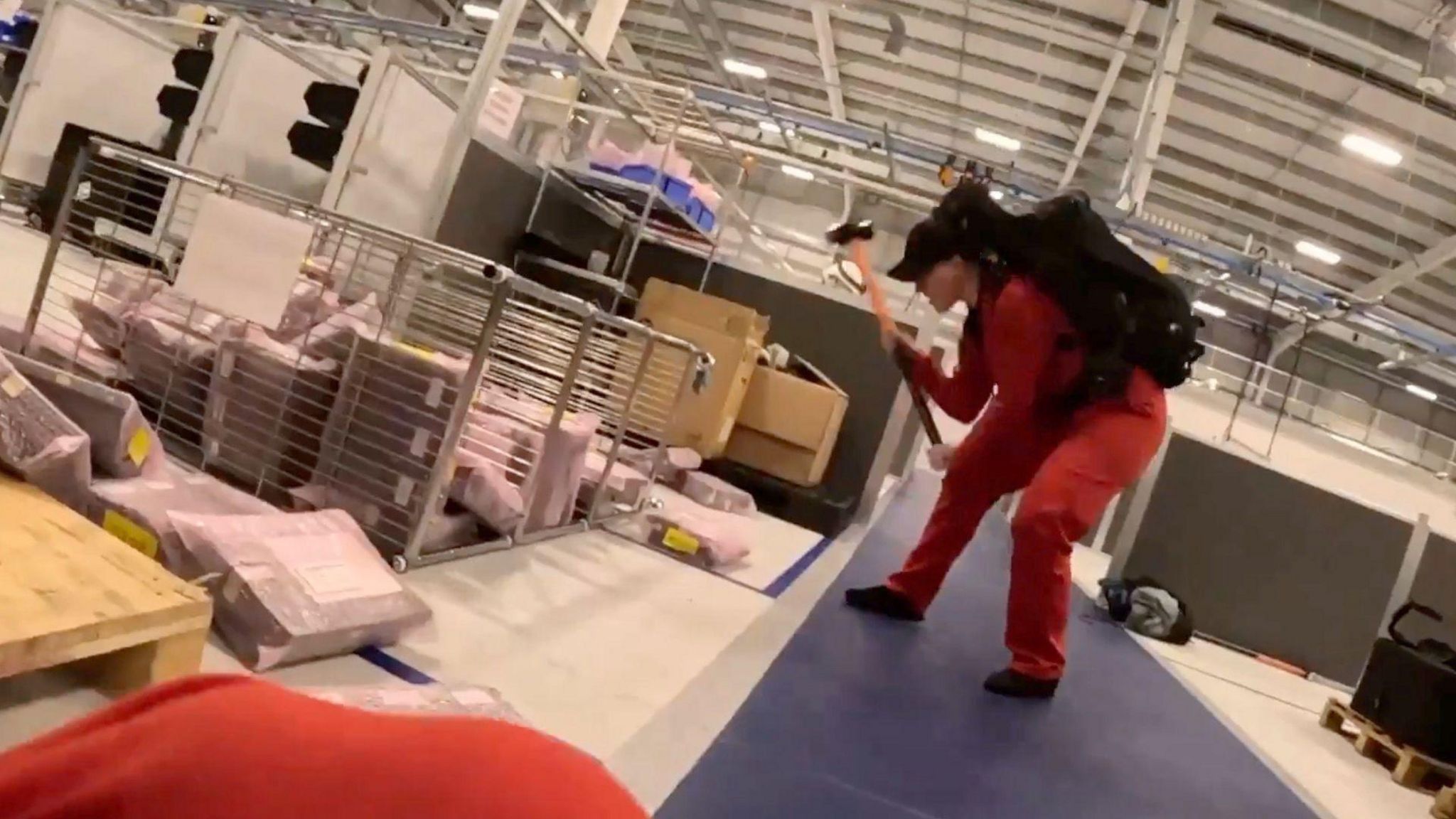 A screenshot of the video shared by Palestine Action, showing a person in a red jumpsuit with a large black backpack on and a black cap, using a sledgehammer to smash equipment in a warehouse. 