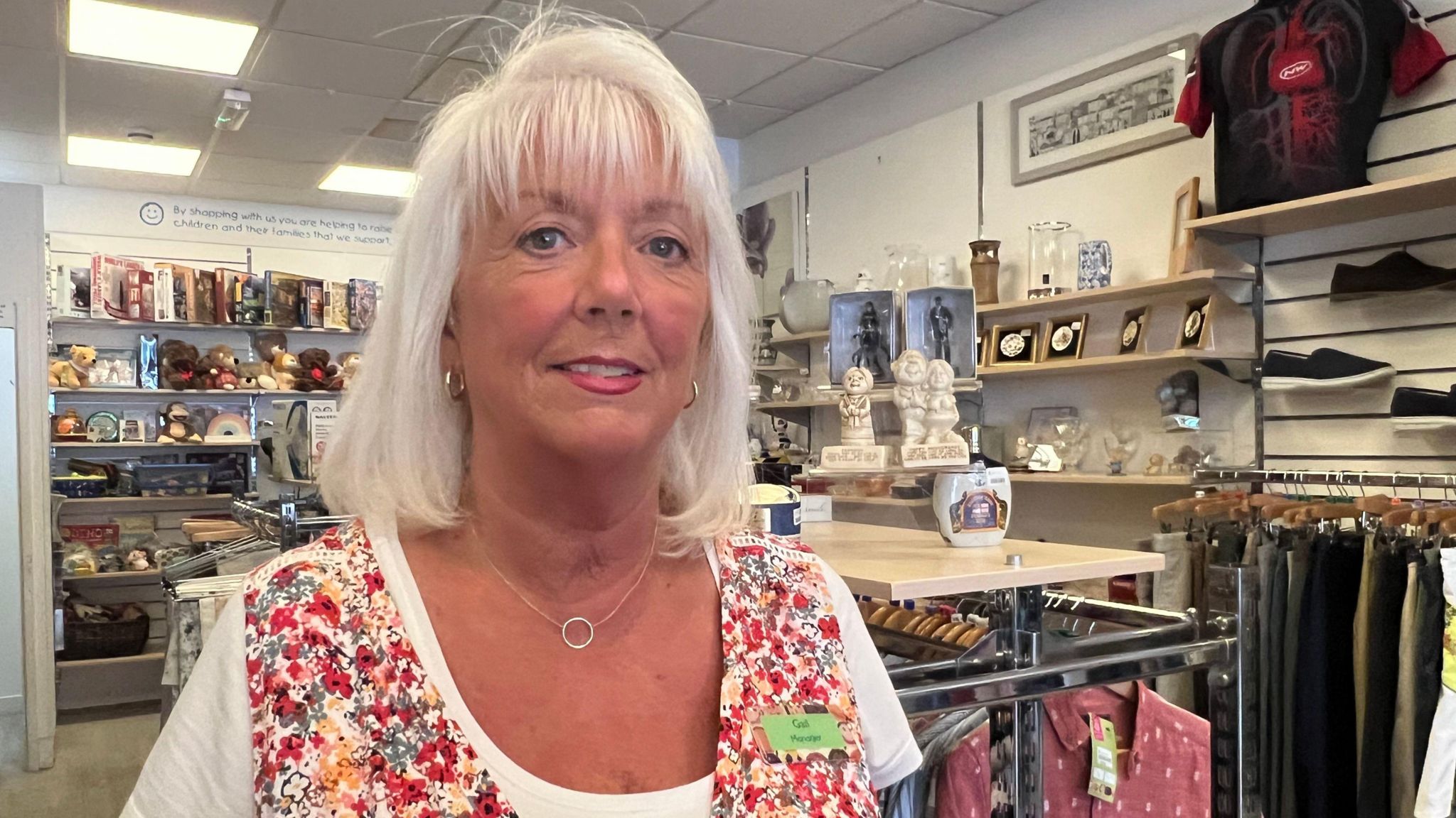 Gail Fry inside a charity shop. She wears a white T-shirt and a floral vest, and has white hair cut into a bob with a fringe. 