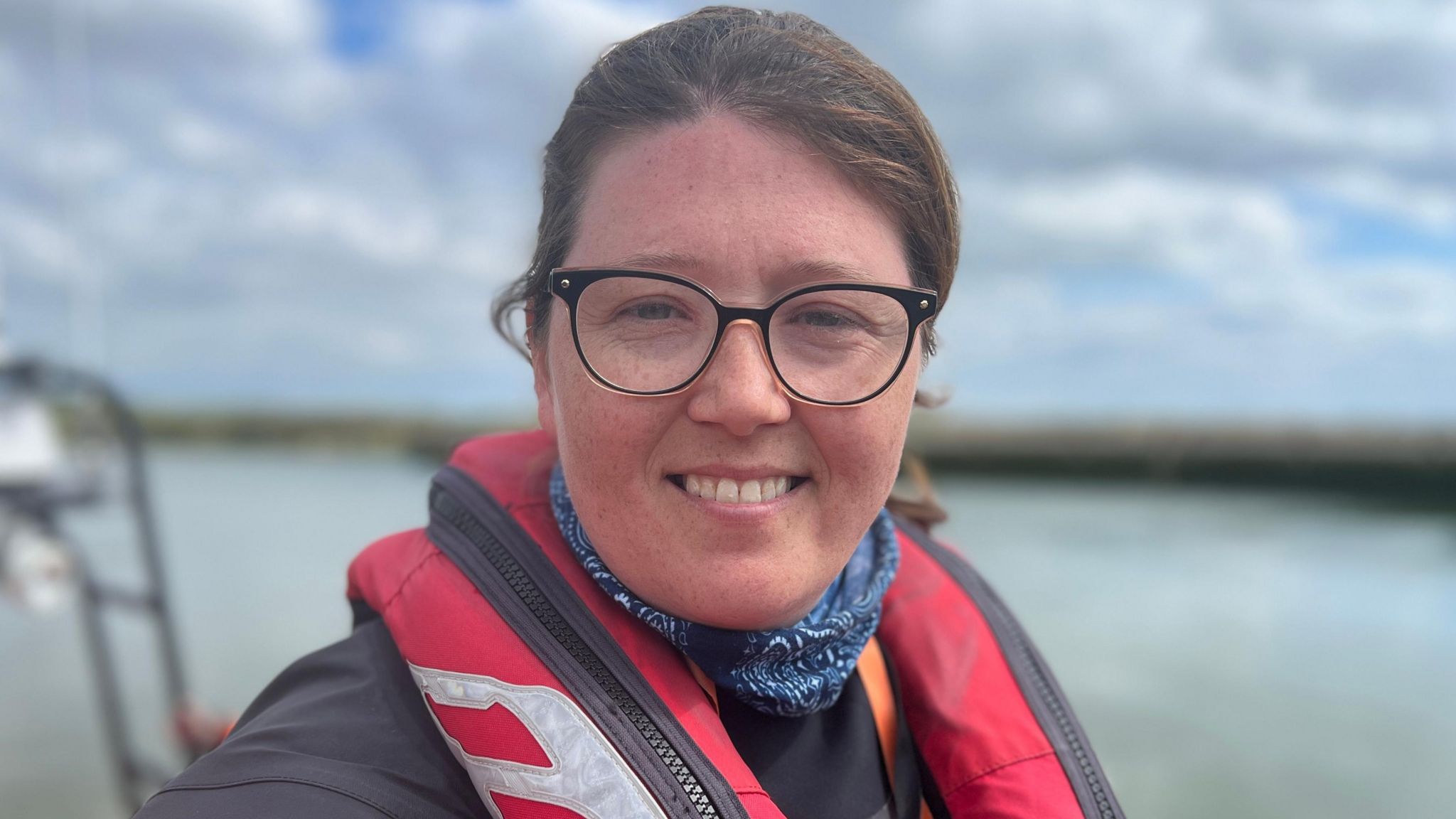 A smiling Marie Worley on board a lifeboat with the sea behind her. She is wearing a blue jacket and red life jacket with a blue scarf around her neck and glasses. 