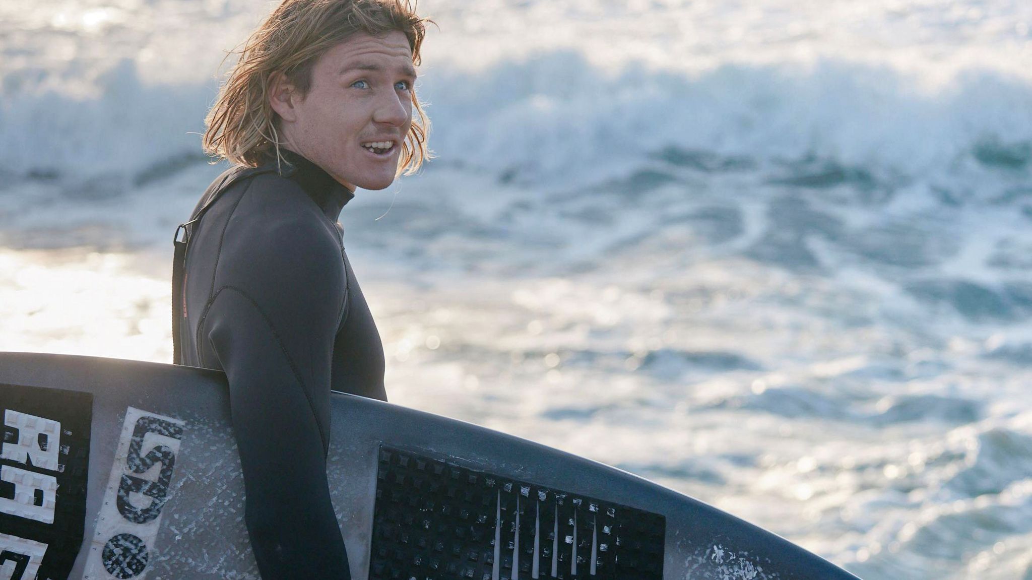 Kai McKenzie, a young man with medium length blonde hair looks on while carrying his surfboard under his arm on the beach with the sea in the background