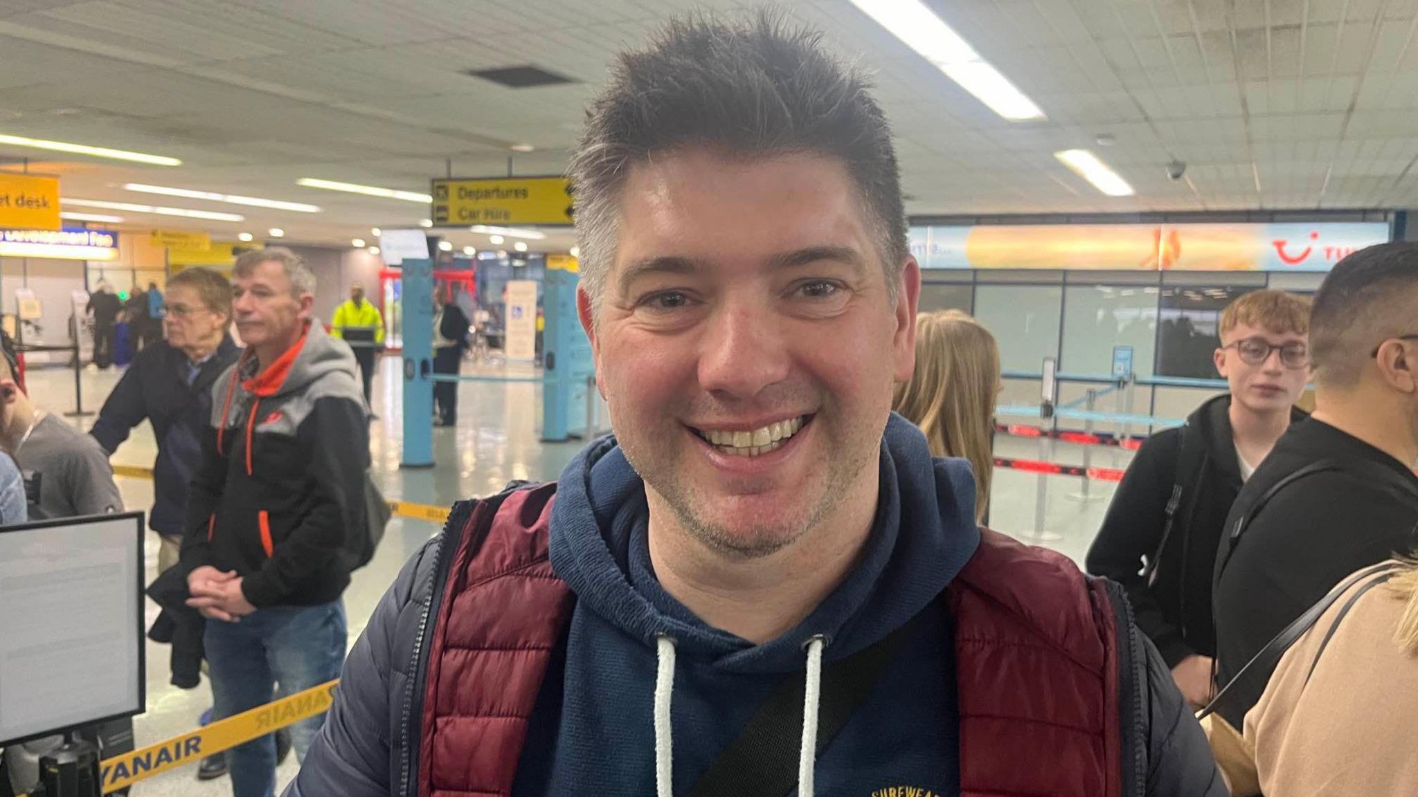 A man smiling at the camera while at Norwich Airport. He is wearing a navy hoodie and a navy puffer jacket
