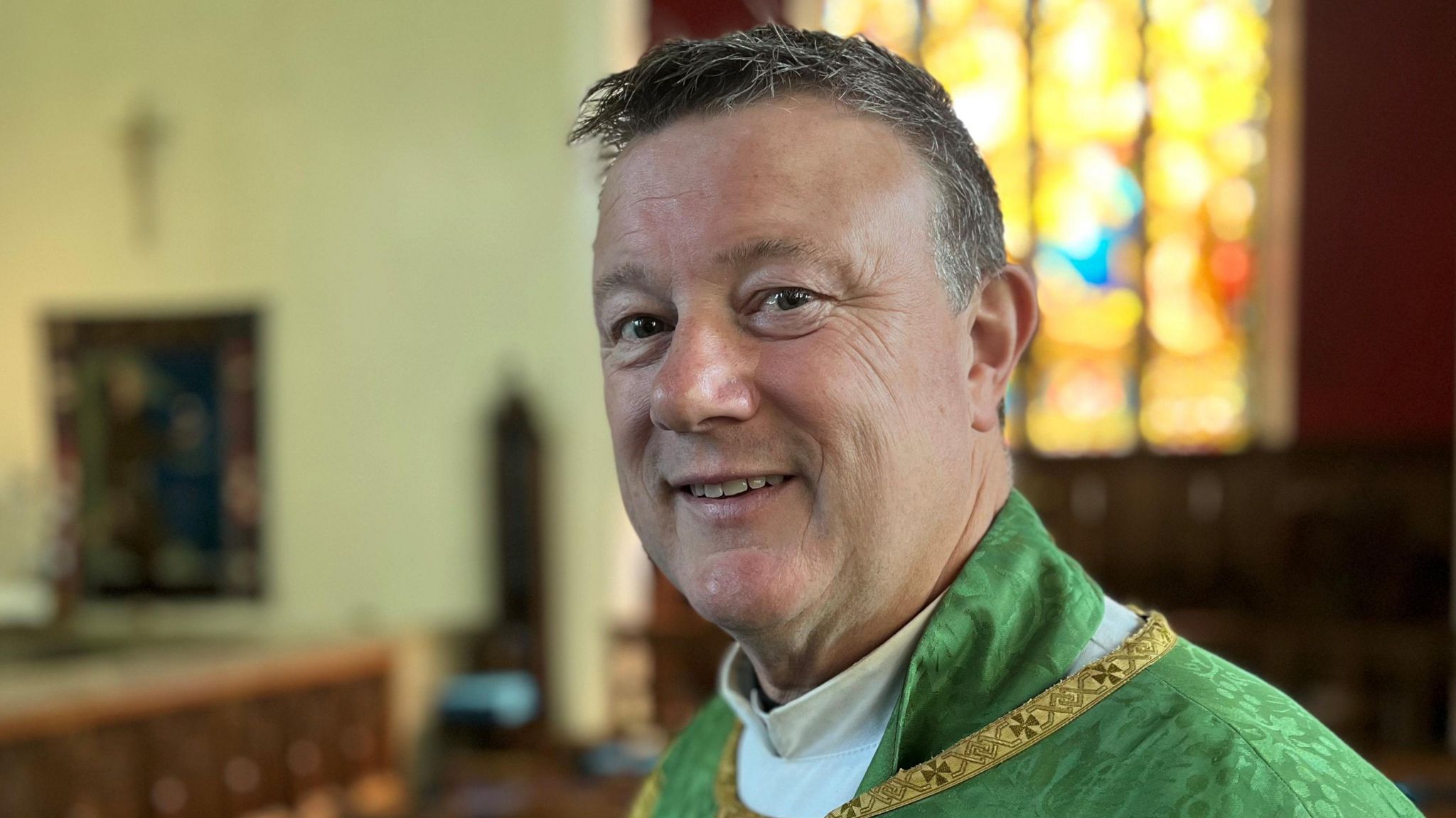 A man in a church wearing green vicar's robes, with grey hair smiling at the camera