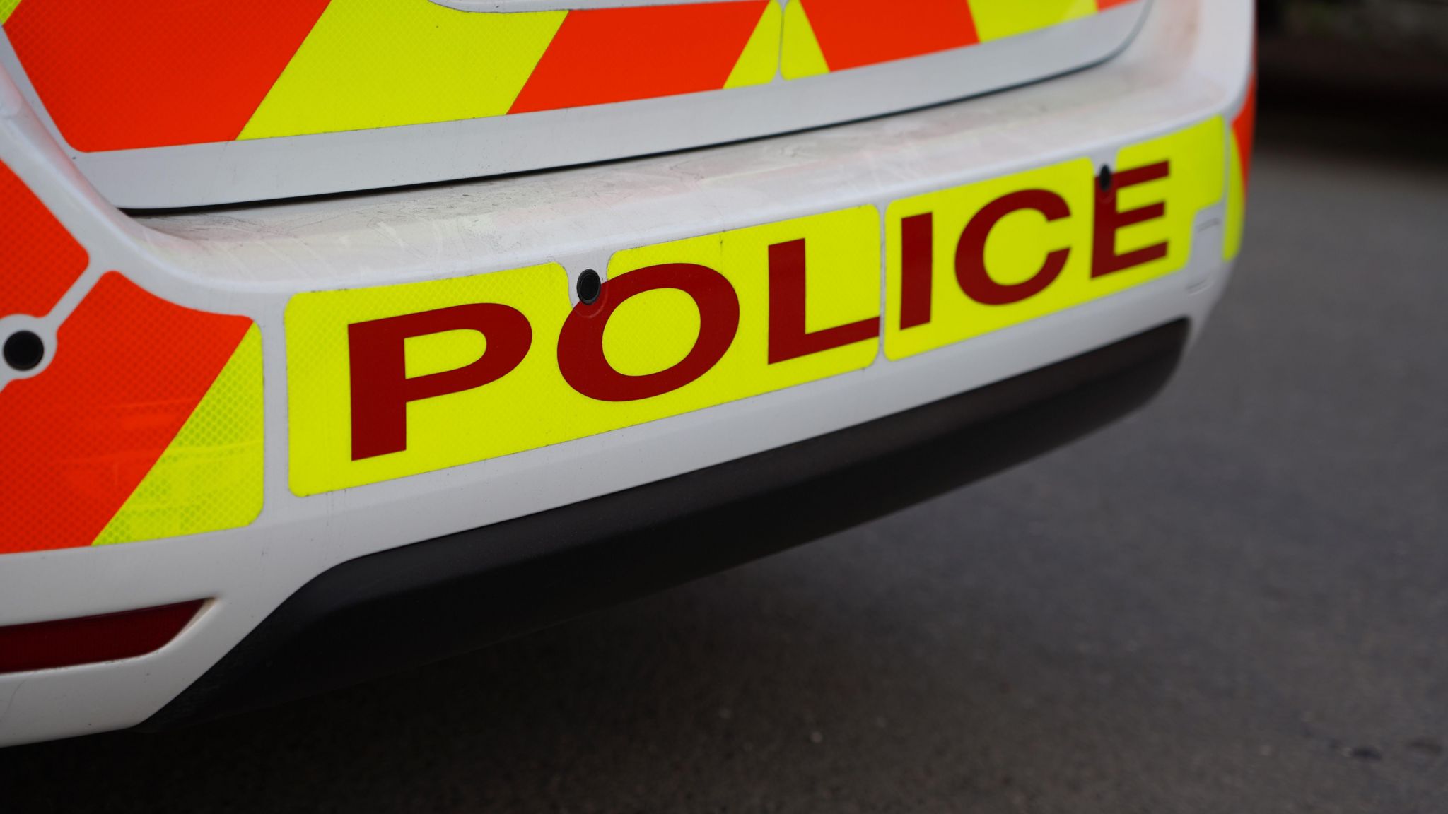 Police car. The bumper of the car is in the shot. It is printed with yellow and orange stripes and police in capital letters along the bottom. 