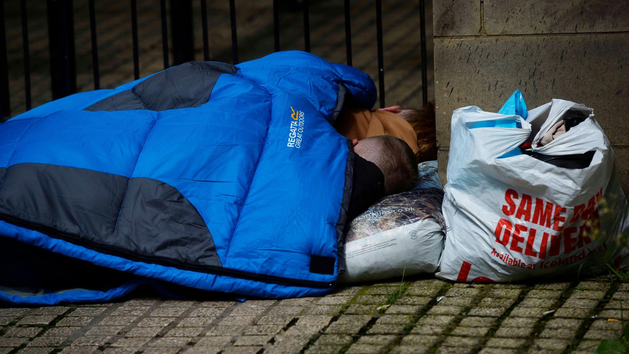 The backs of two people sleeping in a blue sleeping bag on the ground, with their possessions in plastic bags surrounding them