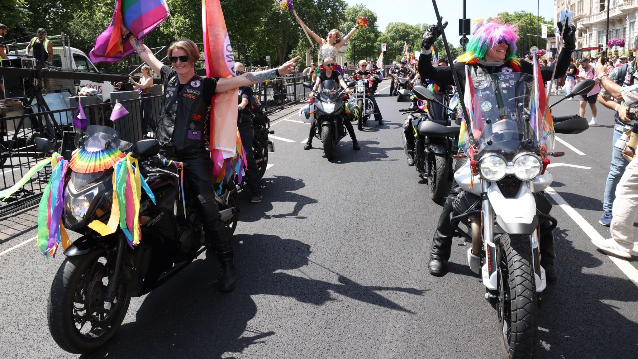 Two people sit on motorbikes decorated with ribbons and Pride flags
