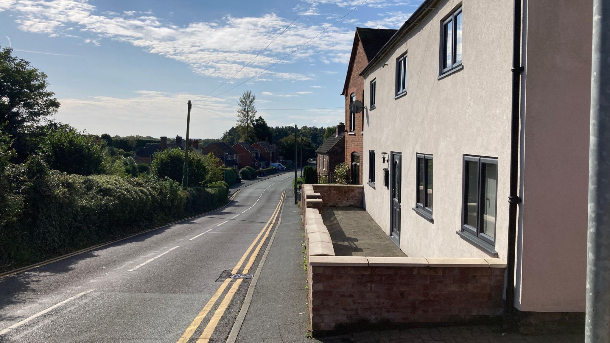 A road with two houses on the right hand side. On the left hand side are trees and bushes