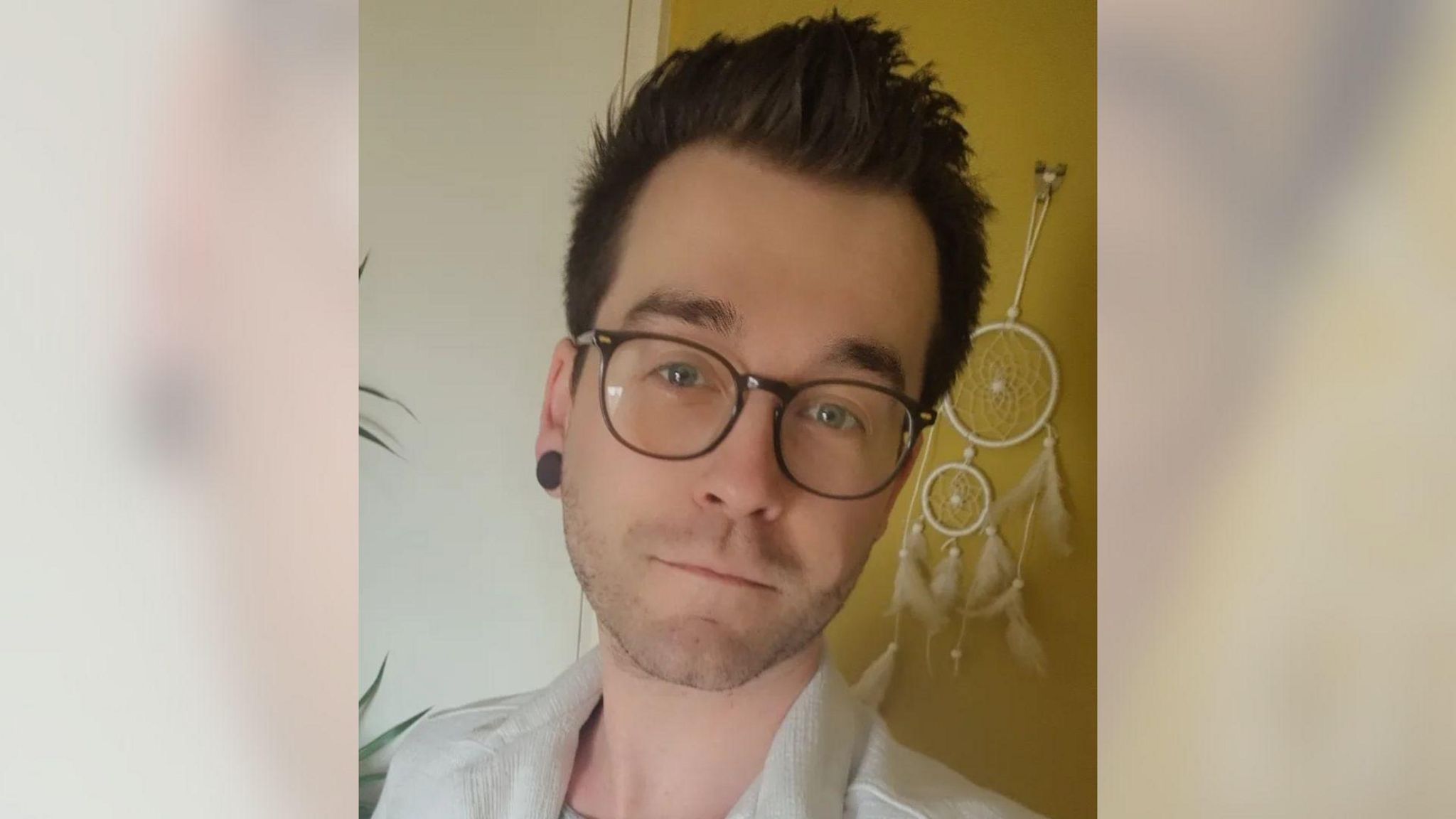 A white man with brown hair and short stubble smiling at the camera. He is wearing brown round glasses and a white shirt. He is stood in front of a yellow and white wall