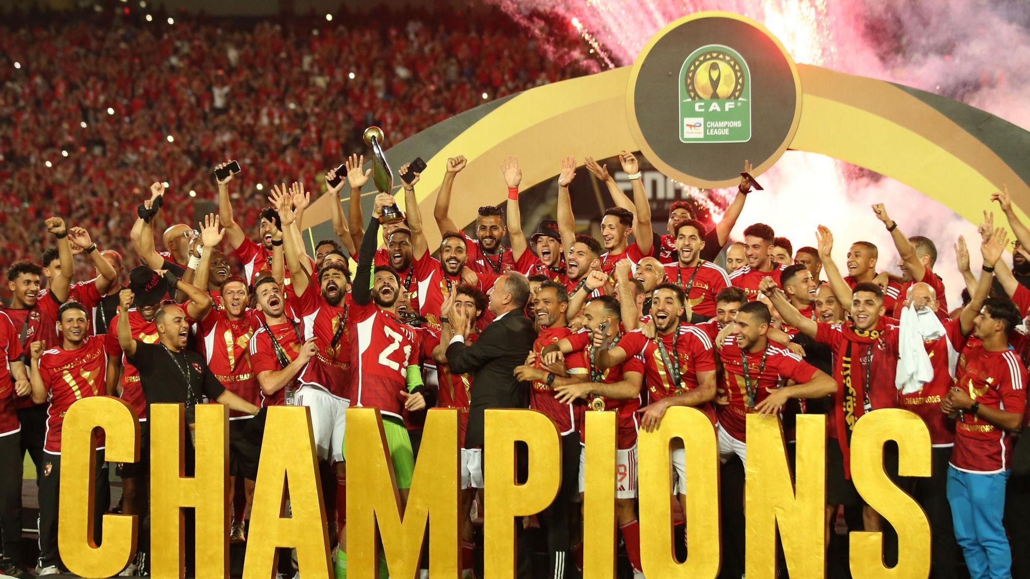Egyptian team Al Ahly celebrate lifting the trophy for the 2024 Caf Champions League, wearing red shirts and standing behind a huge gold lettering that reads "champions"