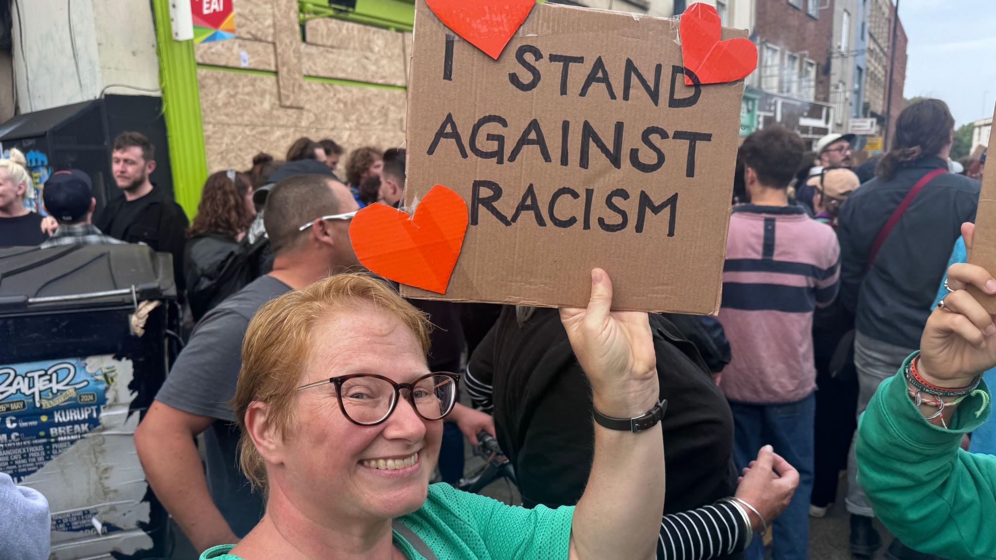 A woman holding a sign reading "I stand against racism". It has hearts on it. She is smiling at the camera. 