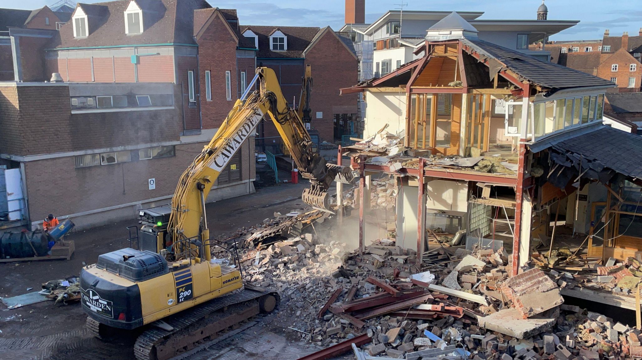 A large yellow digger with a claw is pointing towards a half-demolished building. There are bricks, rubble and wood all over the floor.