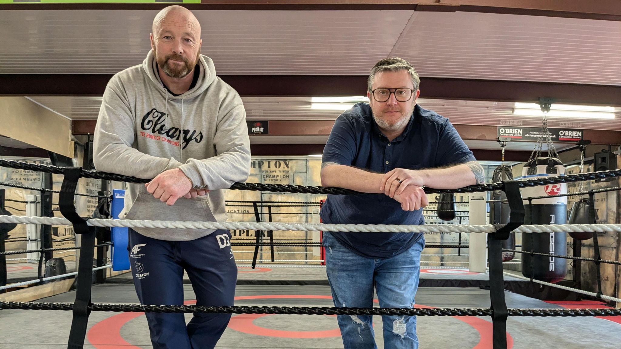 Edwin Cleary wearing a grey hoodie and Kurt Canavan wearing a blue shirt and glasses, they are standing in a boxing ring and leaning on the ropes