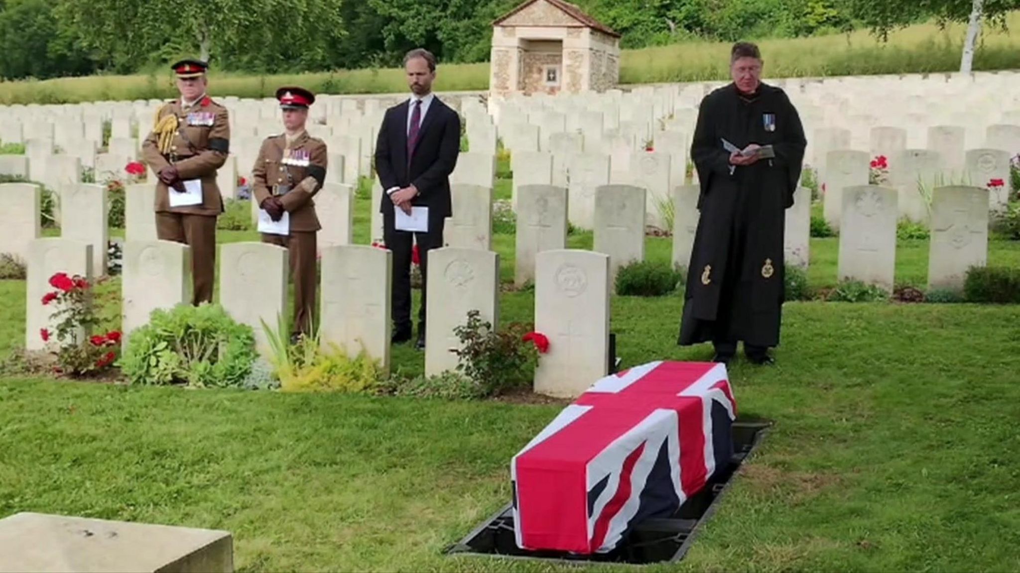 A funeral service at a British war cemetery in France, a coffin with a British flag draped across it is being lowered into the ground and a vicar is reading beside military personnel 