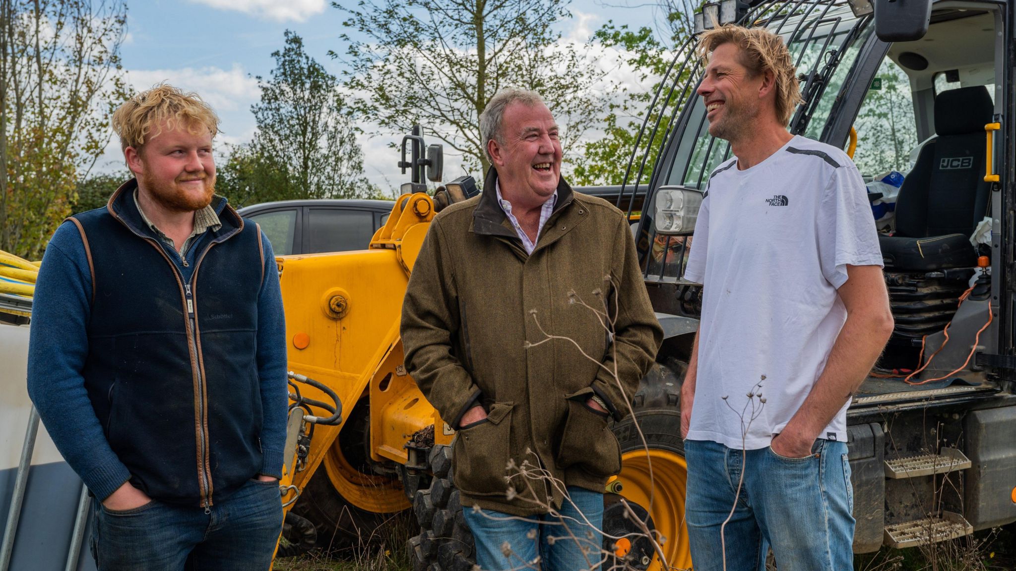 Andy Cato laughing with Jeremy Clarkson and Kaleb Cooper on Diddly Squat farm in the Cotswolds, standing in front of a telehandler.