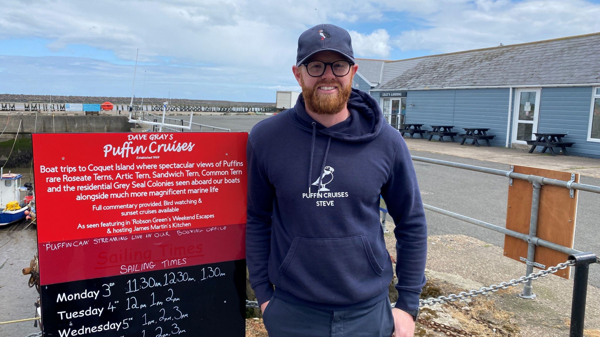 Steven Banks standing in front of a sign saying Puffin Cruises in Amble harbour 
