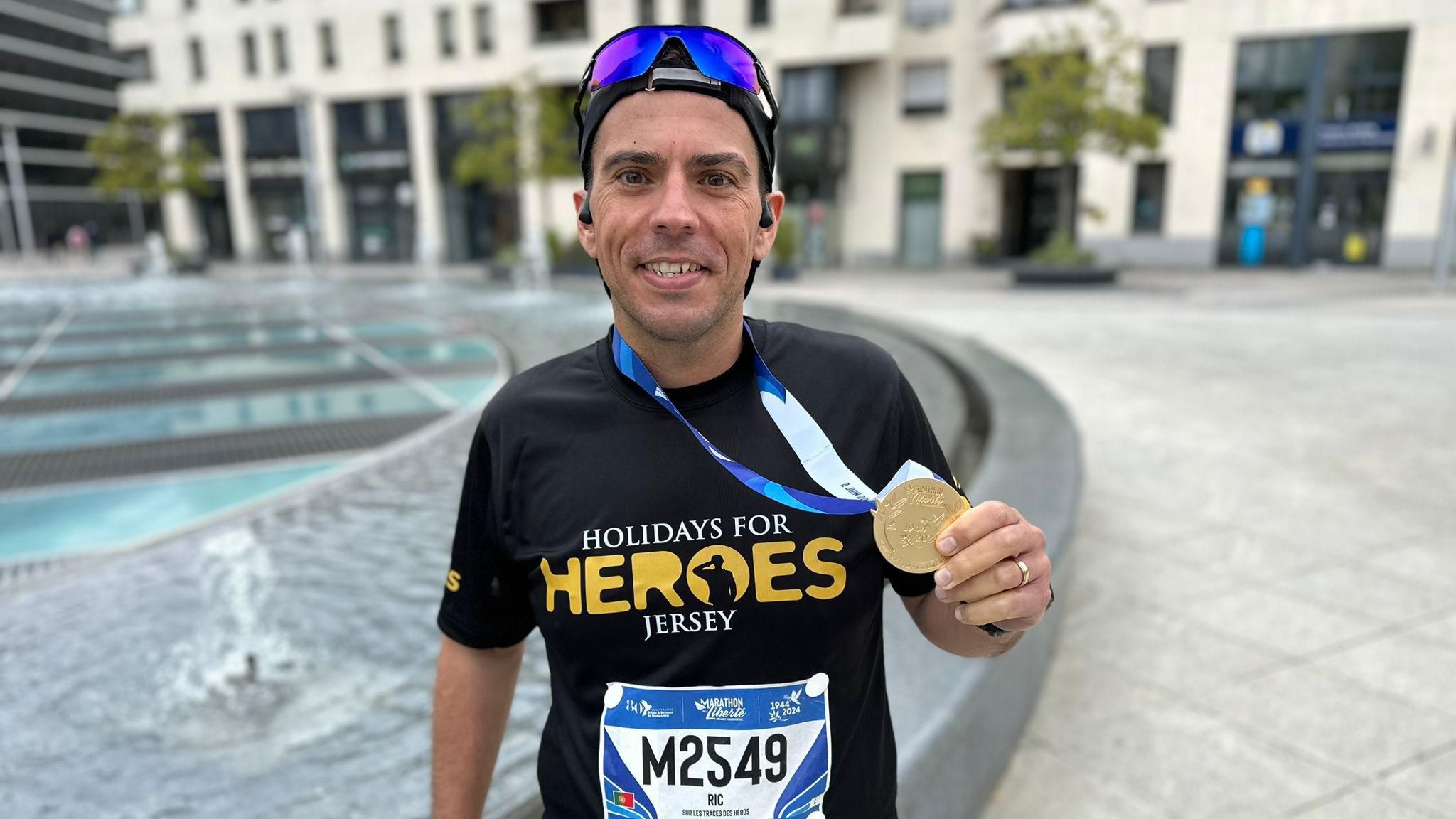Ricardo smiles at the camera with his medal around his neck and he is wearing a Holidays for Heroes Jersey running top