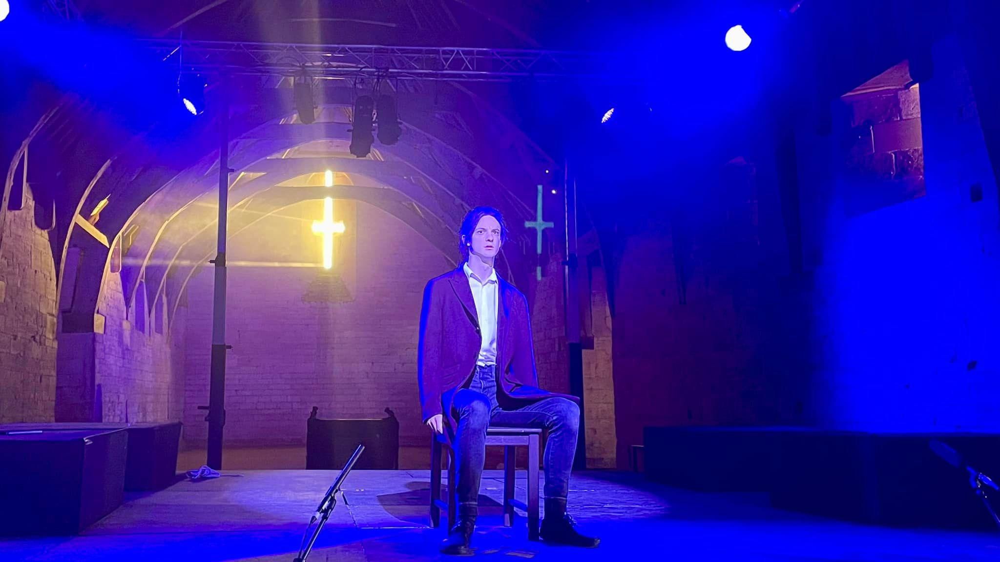 A man wearing jeans and a blazer sitting on a wooden chair in the middle of the stage at Tithe Barn. It is dark and there is a blue smoky light filling the room