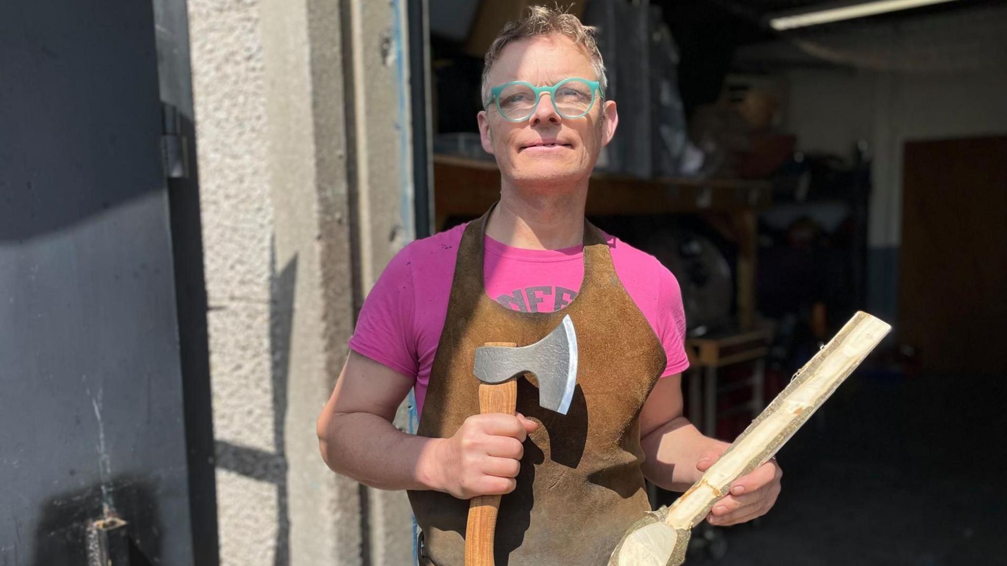 A man with green glasses and brown apron is holding an axe and a large wooden spoon