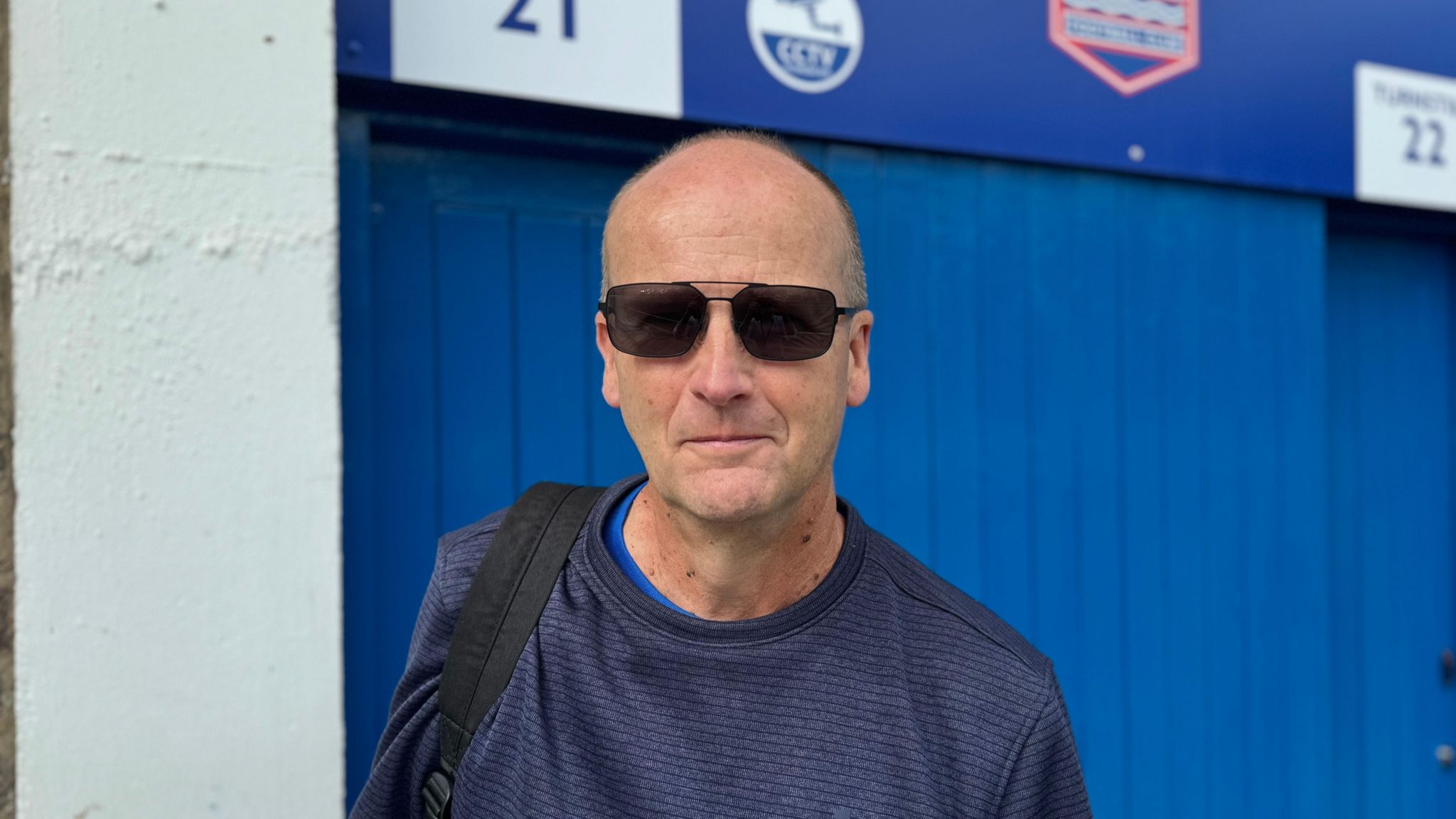 Ipswich Town fan David Grey, wearing sunglasses and a blue jumper, standing in front of the club's Portman Road stadium