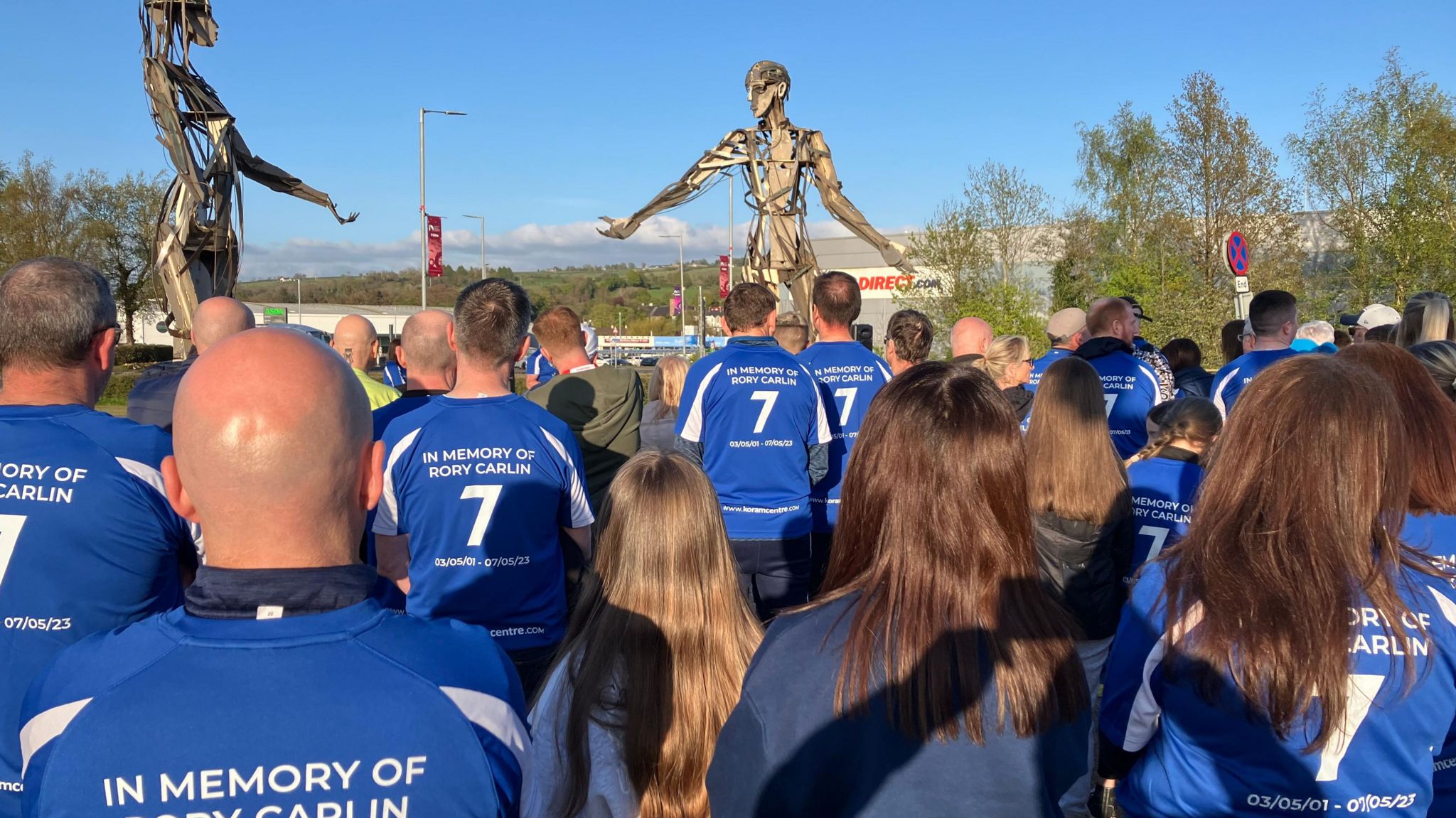 Dozens of people with their backs to the camera, wearing football shirts that say "In memory of Rory Carlin" on the back