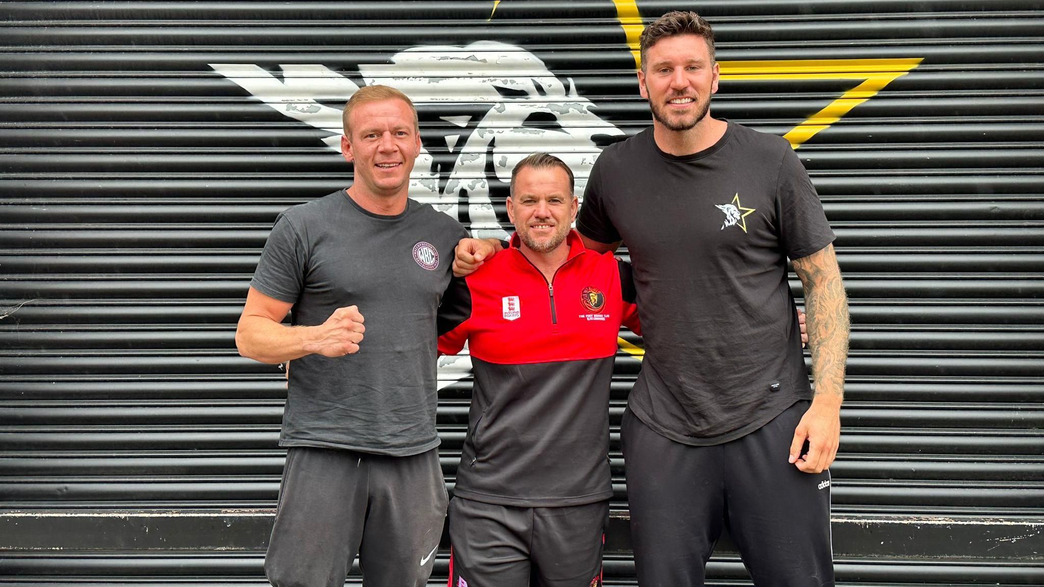 Three men are standing in front of a black metal shutter. Carl Dickins is on the left. He is wearing a black t-shirt and black shorts and has strawberry blonde hair. Dan Bailey is in the middle, he has brown hair and facial hair, and is wearing a black and red t-shirt and black and red shorts. On the right is Joe Lockley, he is wearing a black t-shirt and black shorts. He has brown hair and facial hair.