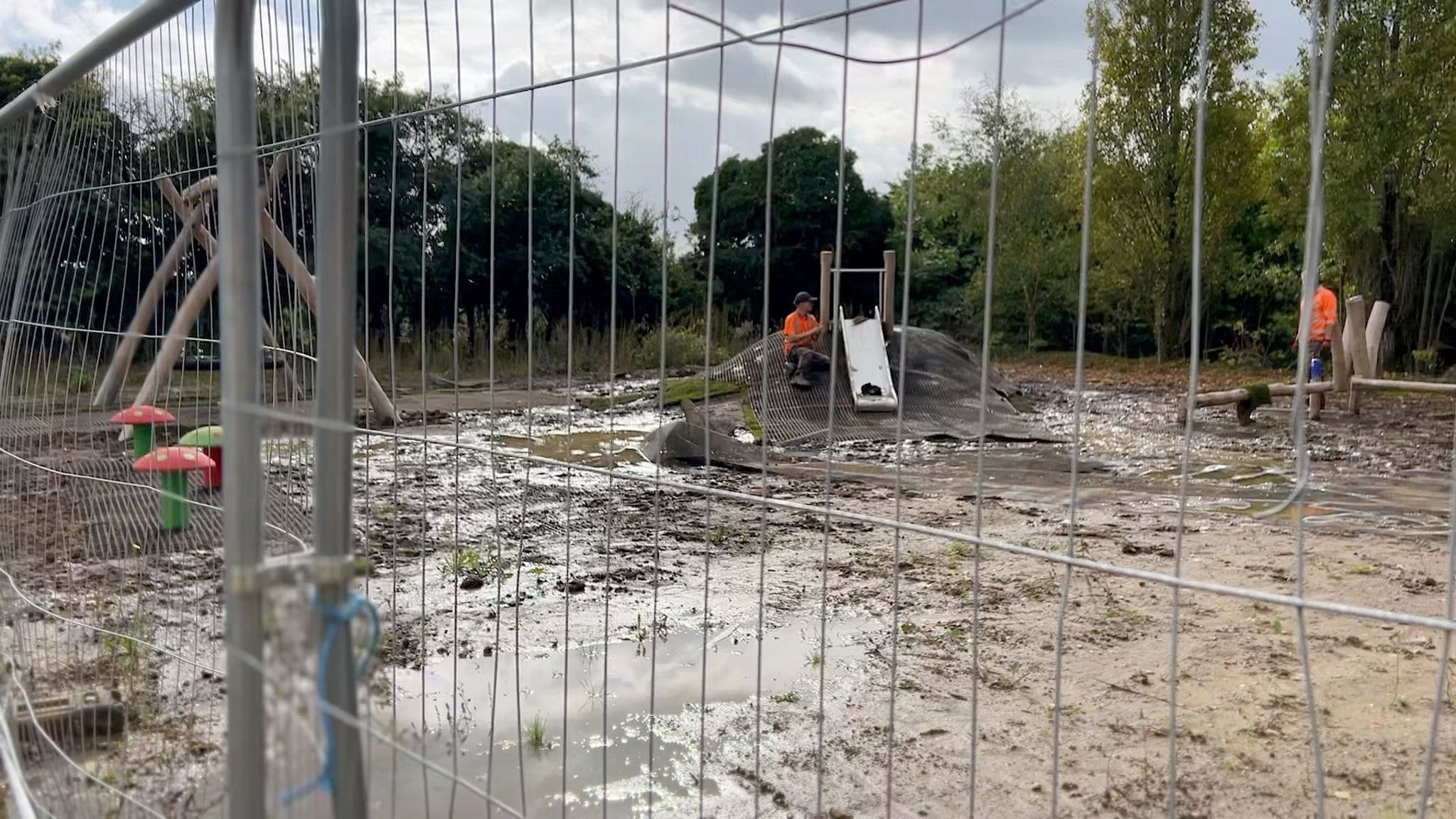 Two workers in orange hi-vis tops in a partially constructed playground. The play equipment is not finished and the ground is very muddy with large puddles. There is metal security fencing running along the front of the site to prevent entry.