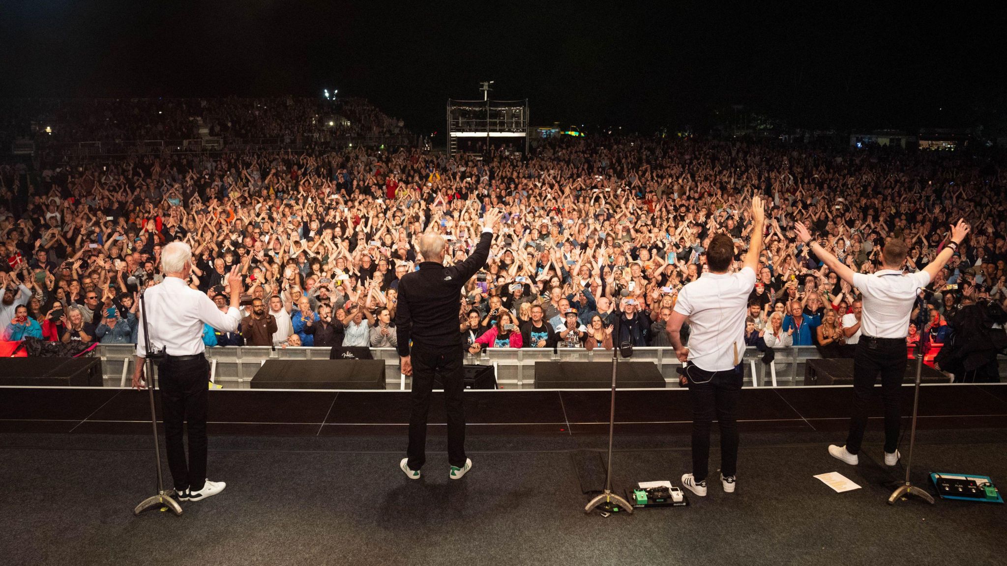 Status Quo standing with the hands in the air facing the audience who also have their hands up and are facing the camera