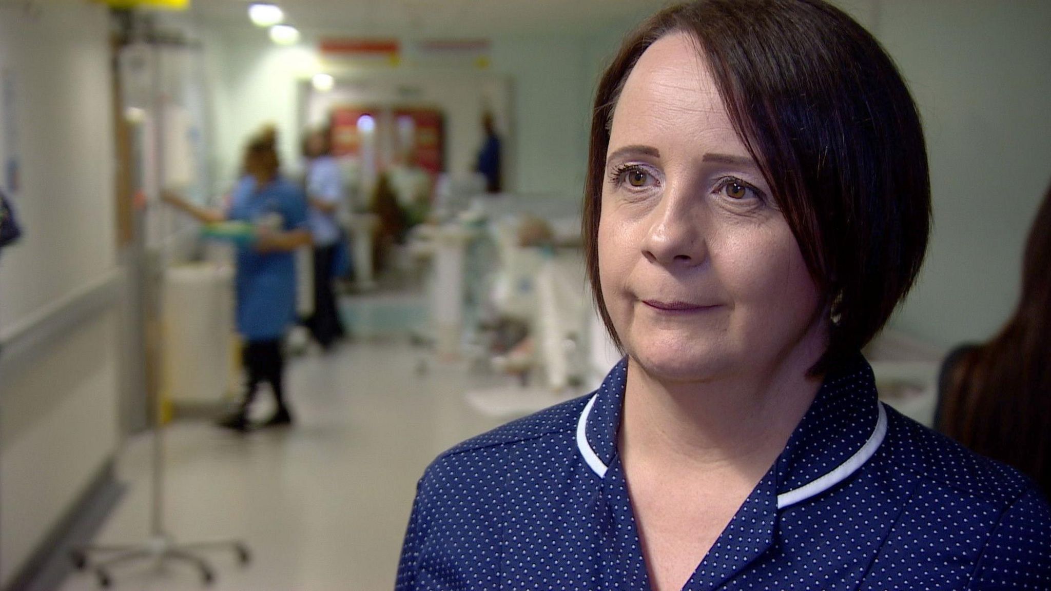 Ruth Green wearing a blue nursing uniform looks off camera . She has dark brown hair in a bob.