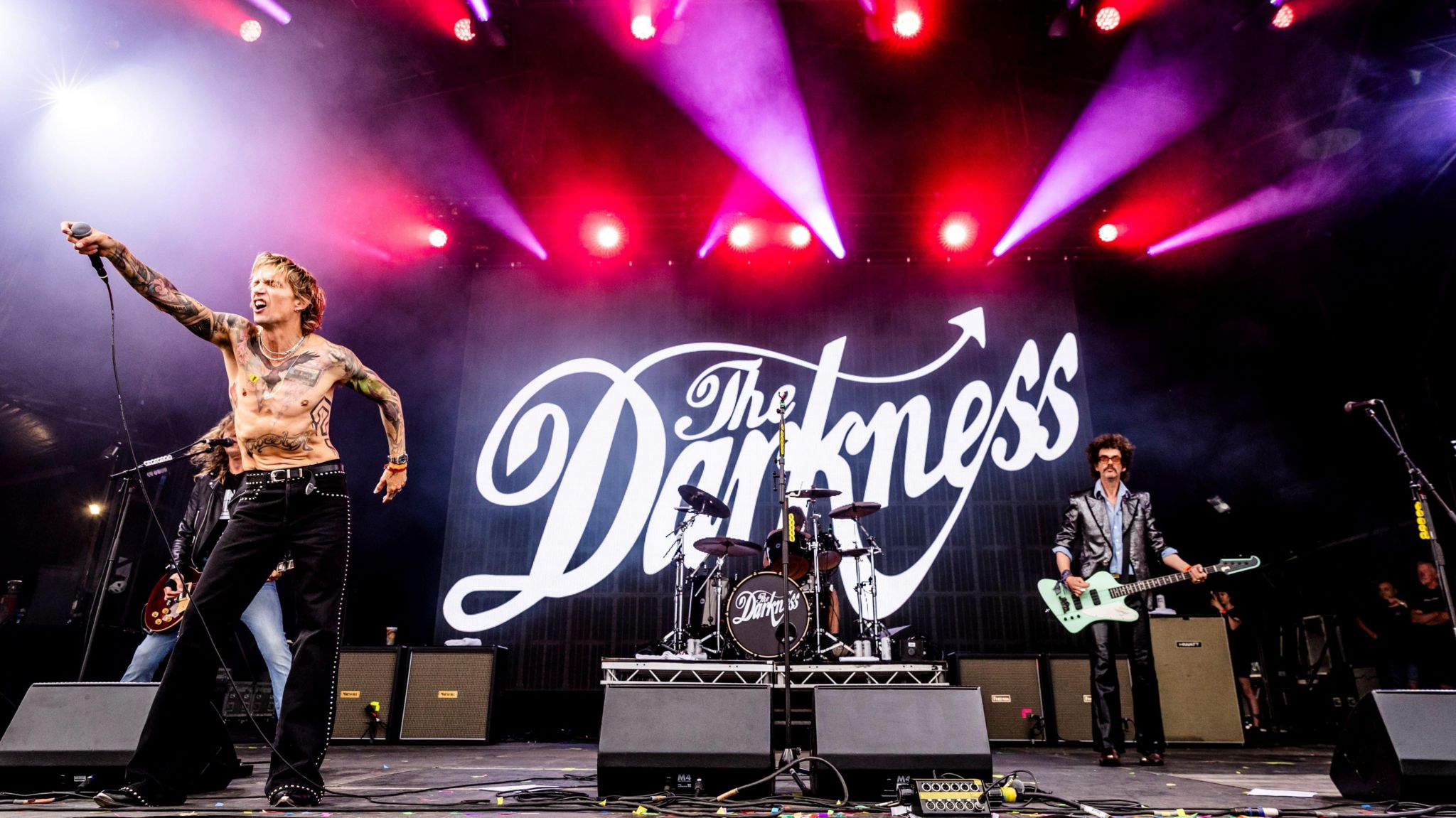 Justin Hawkins, lead singer of The Darkness, holds up a mic pointing at the crowd as the rest of the band performs behind him on stage