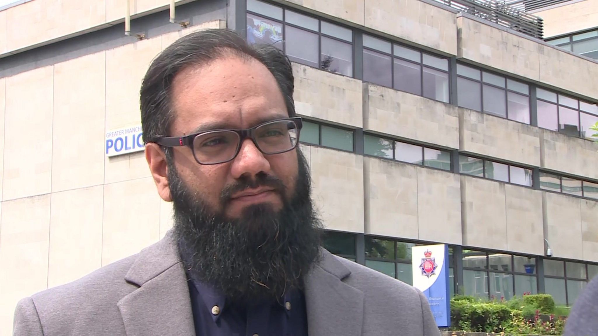 Usman Nawaz from the Rochdale Community Alliance in front of Rochdale Police station wearing a grey blazer and navy shirt. He wears glasses, has dark hair and a mid-length dark beard.