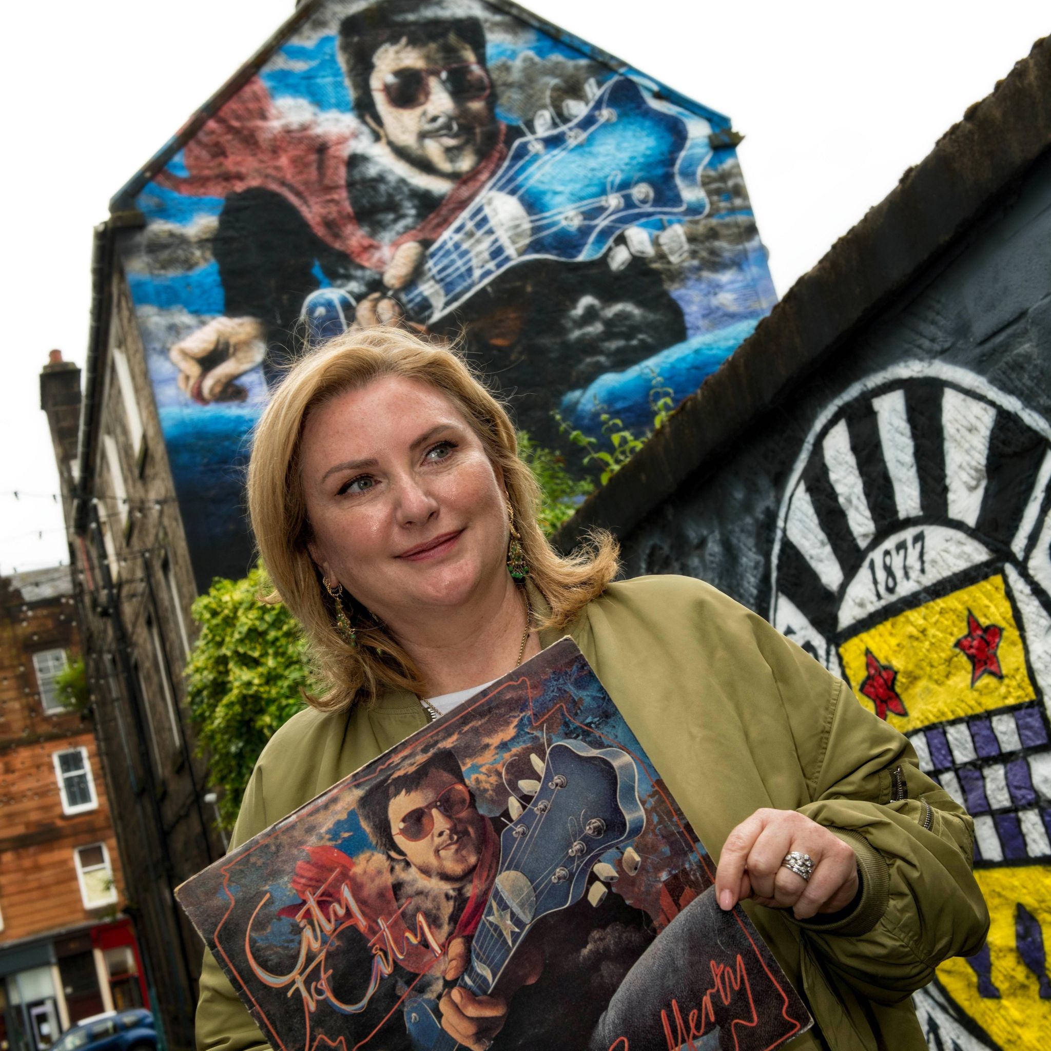 Martha Rafferty holding her dads album cover stood in front of a mural on side of building of same mural