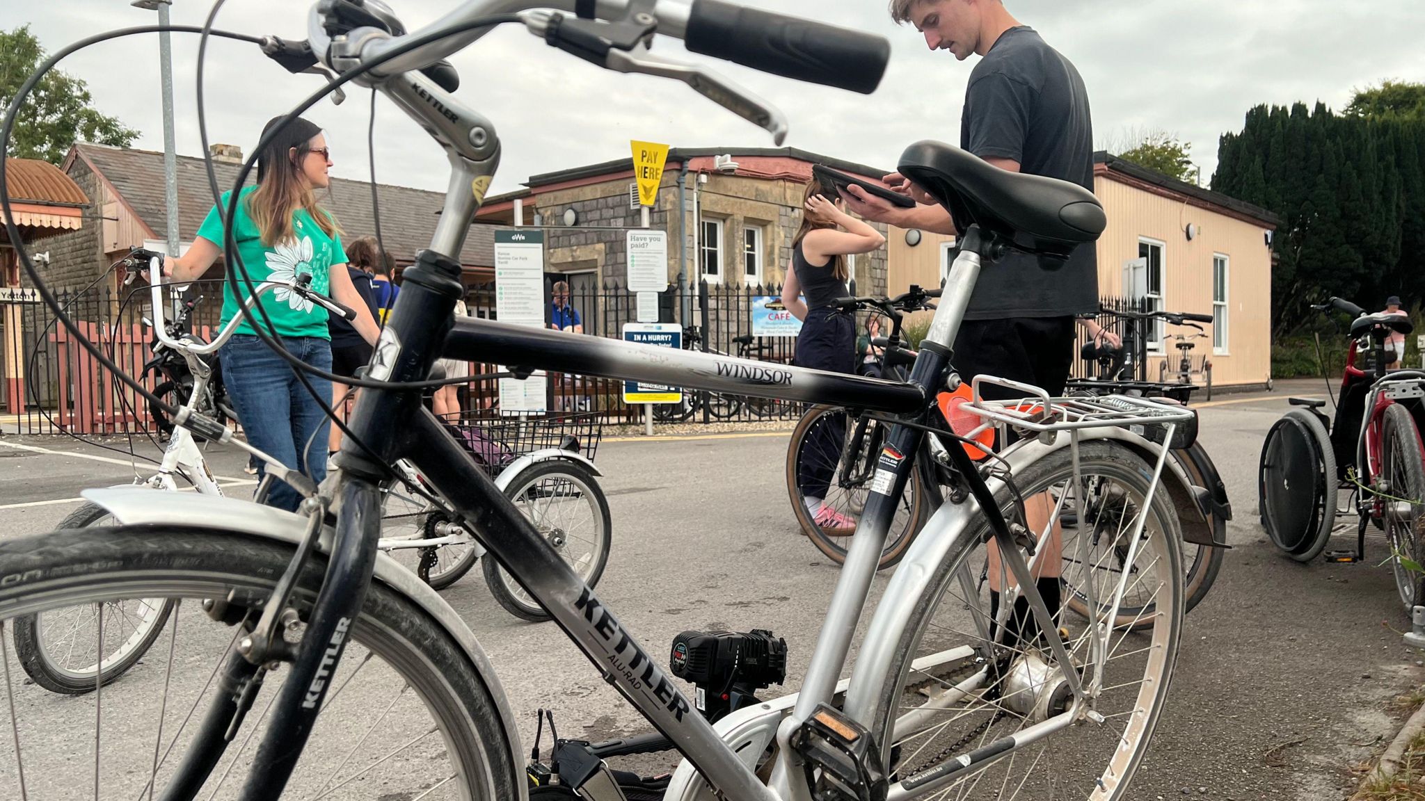 A silver bike is visible in the foreground, in front of a train station. There are also people visible in the background, all looking away.