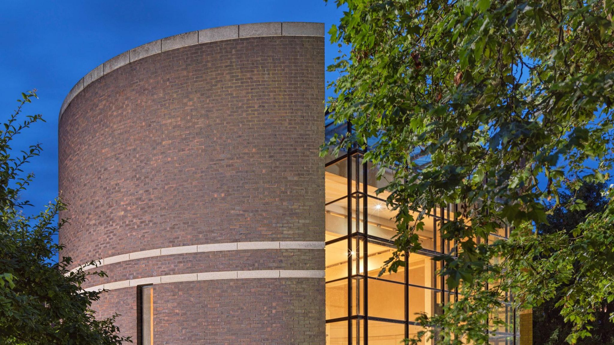 Fitzwilliam College's chapel at dusk, a brick and glass building with a tree in the foreground 