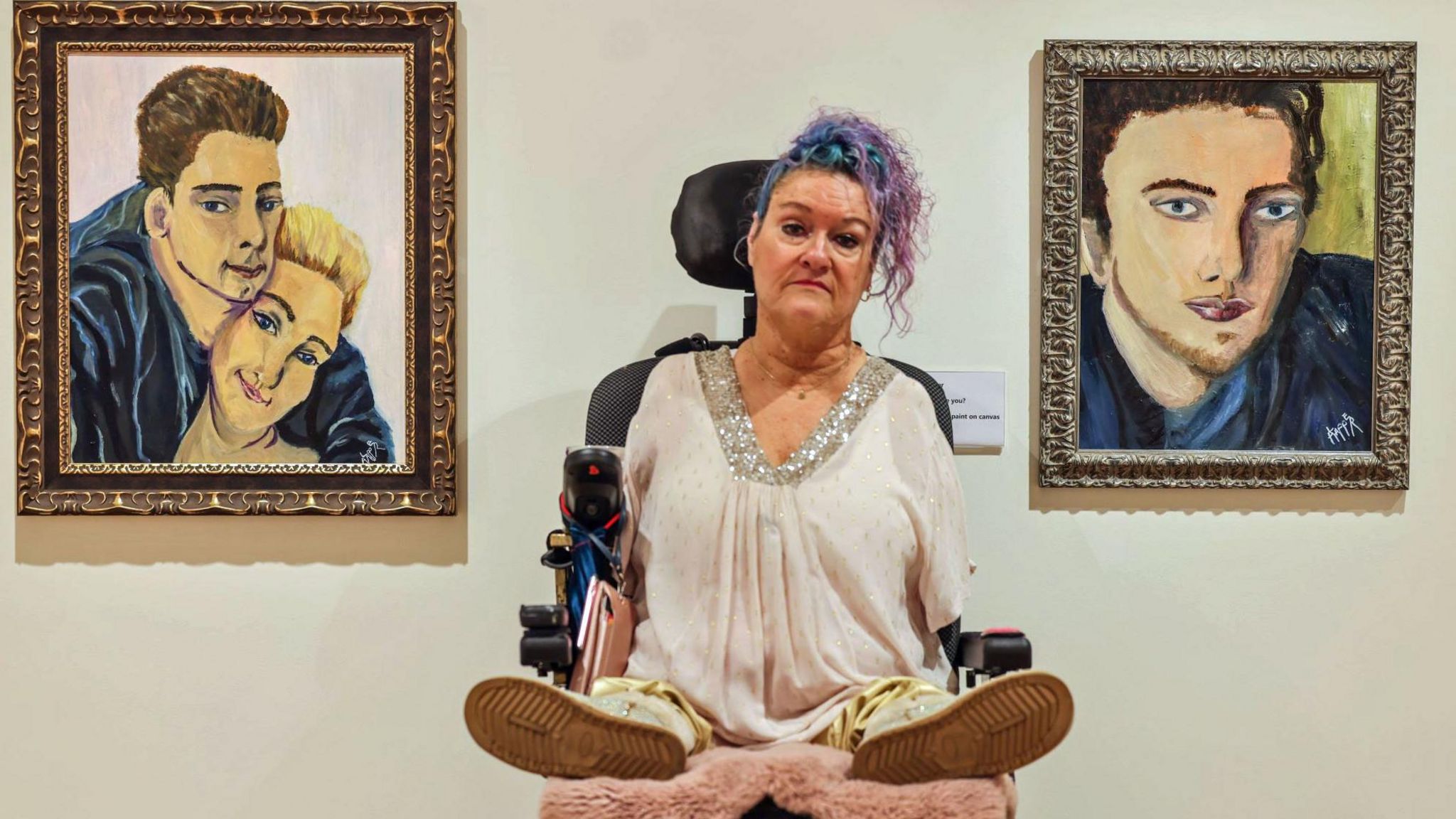 Alison Lapper sits in her wheelchair besides two portraits she painted of her son Parys. The portrait on the left is a painting of Parys cuddling Alison. They are smiling. The portrait on the right is only of Parys and his eyes are widened with a pensive look on his face. 