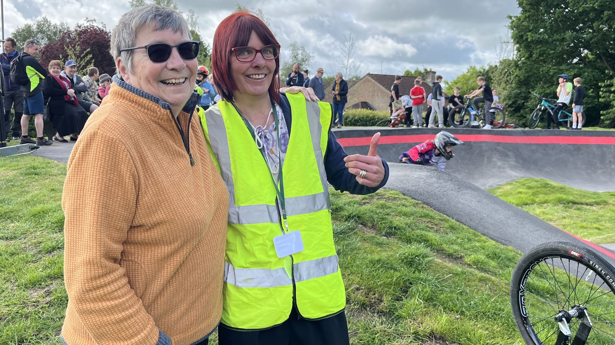 Pewsey Parish Councillors, Marilyn Hunt and Lisa Brindley stood on the edge of the pump track, Lisa with a thumb up wearing a high visibility vest and Marilyn wearing sunglasses, both smiling