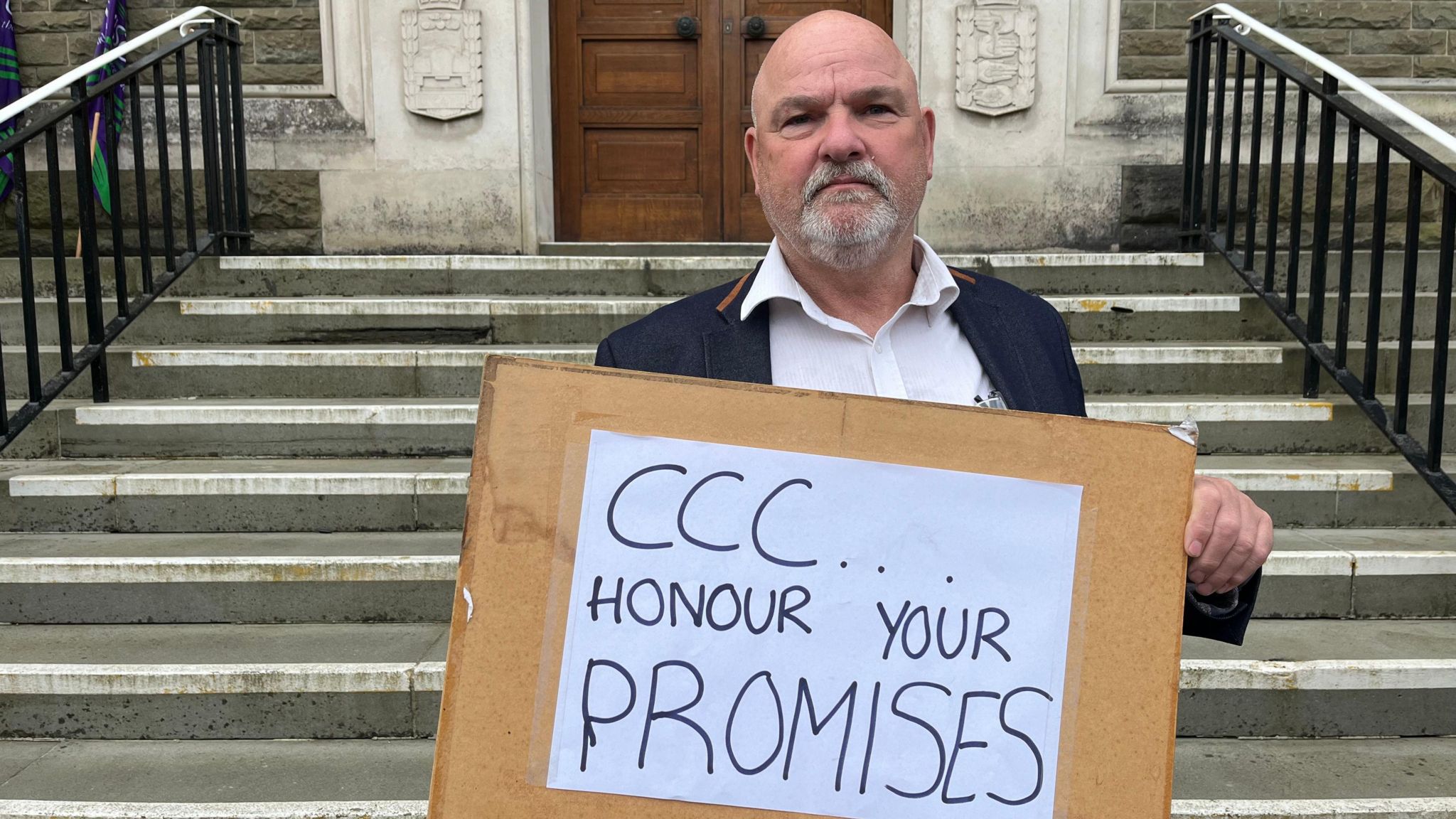 A man stands on the steps of Carmarthenshire County Hall holding a sign that reads 'CCC honour your promises'