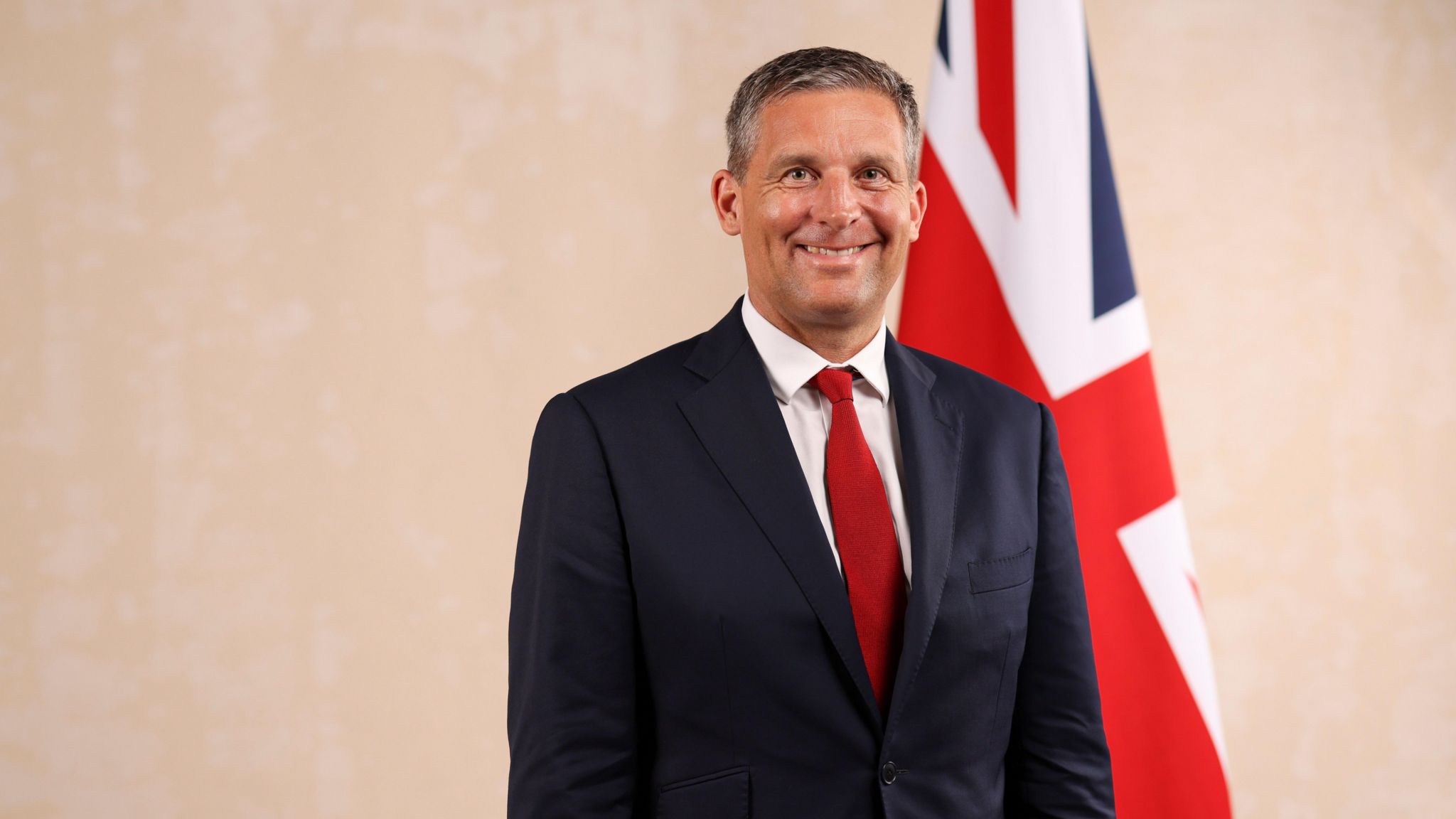 James Timpson wearing a dark navy blue suit, a white shirt and a red tie. He is smiling at the camera