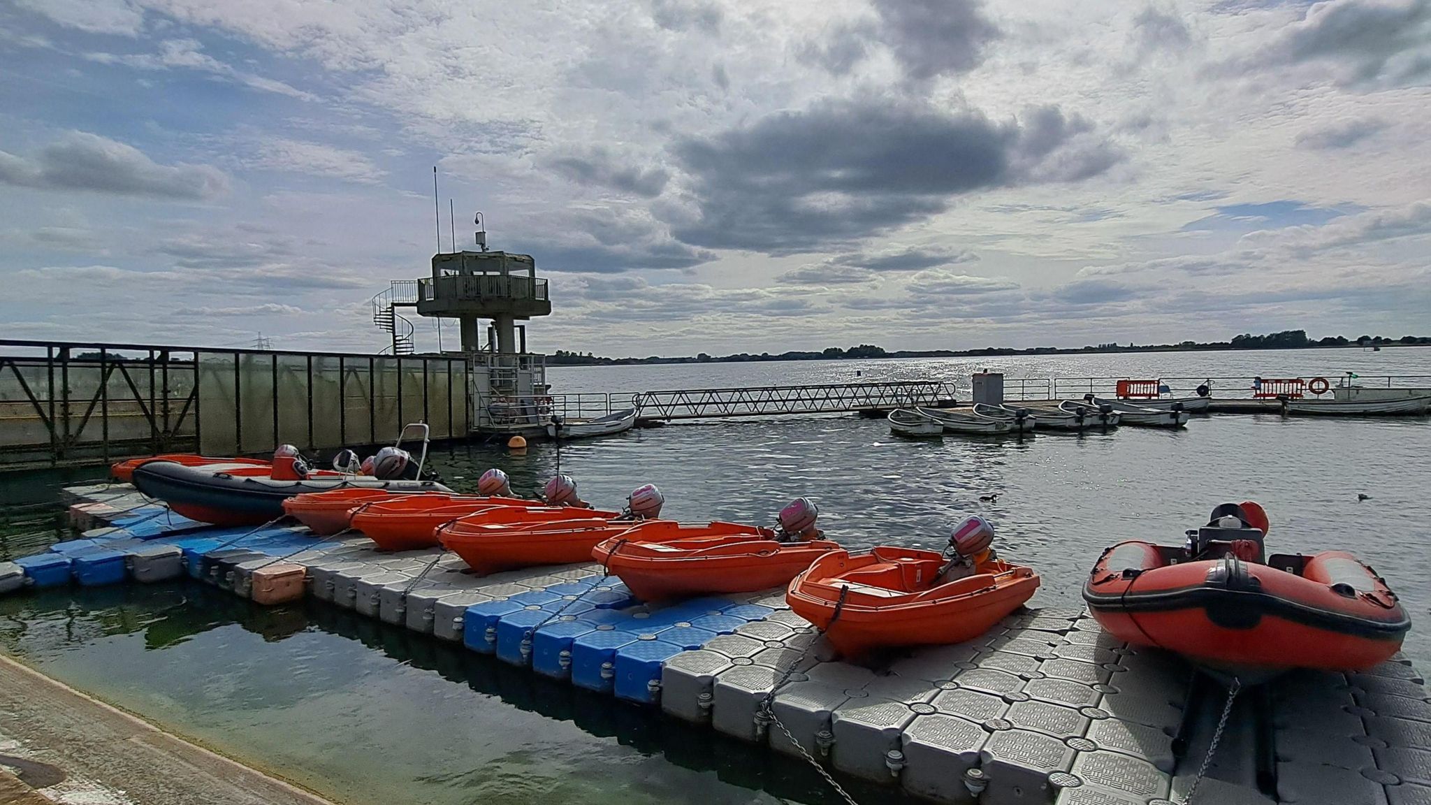 A cloudy bright sky with blue patches gives way to shimmering water of a reservoir. Several orange boats sit on a pontoon in the foreground.